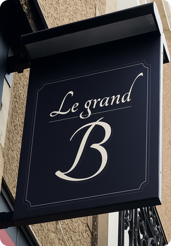 Enseigne à l'extérieur d'un bâtiment fabriquer par LeGraveur signalétique en Martinique avec le texte « Le grand B » écrit en lettres cursives blanches sur fond sombre. La police de caractères est élégante et évoque un style chic et sophistiqué.