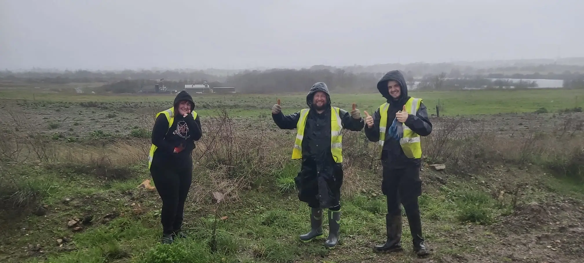Three people in safety gear stand in a field, gesturing towards the horizon under overcast skies.