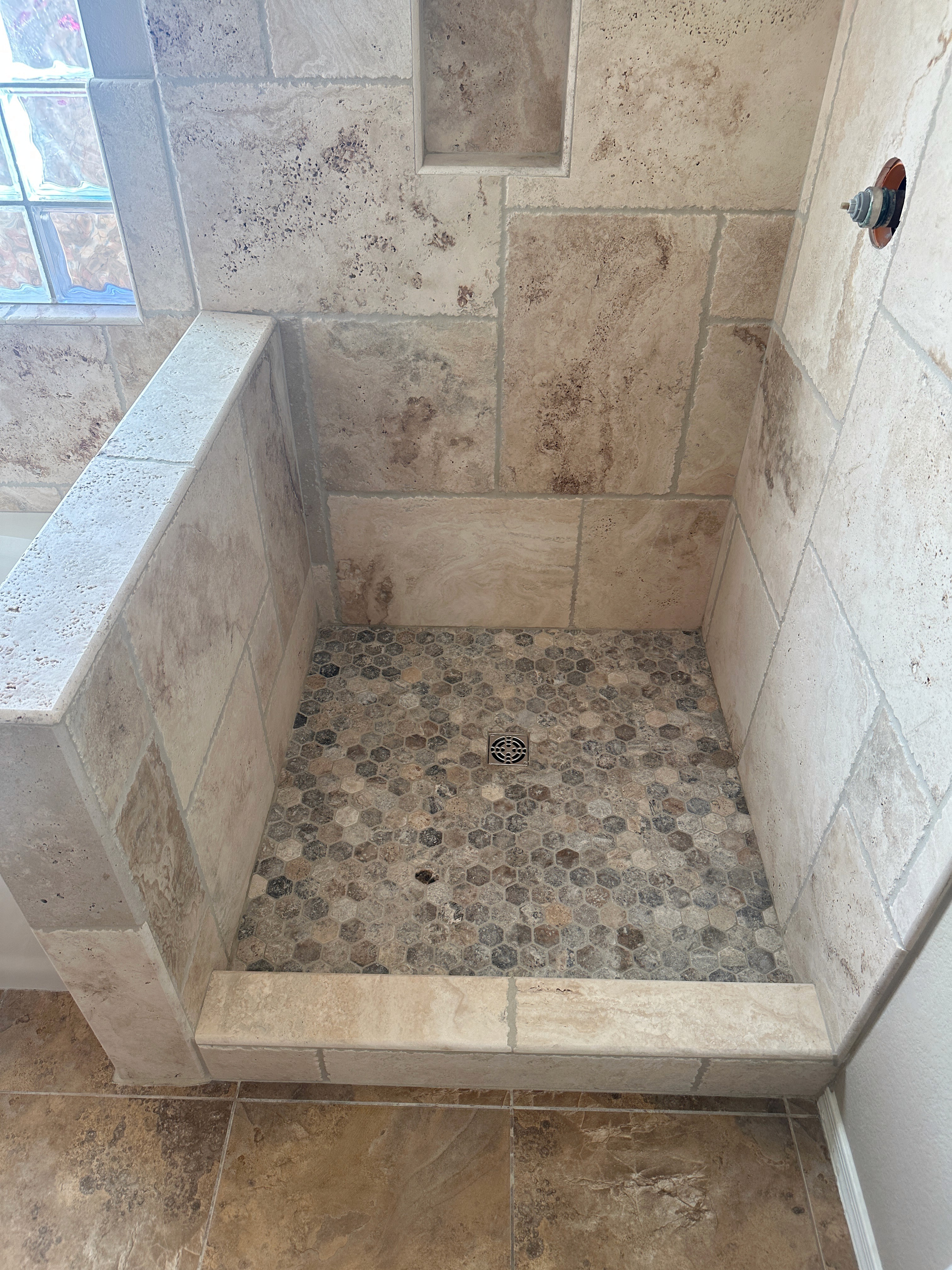 Completed custom shower with light brown natural stone wall tiles and multi-colored pebble mosaic shower floor tiles, featuring a low half-wall and a built-in shower niche.