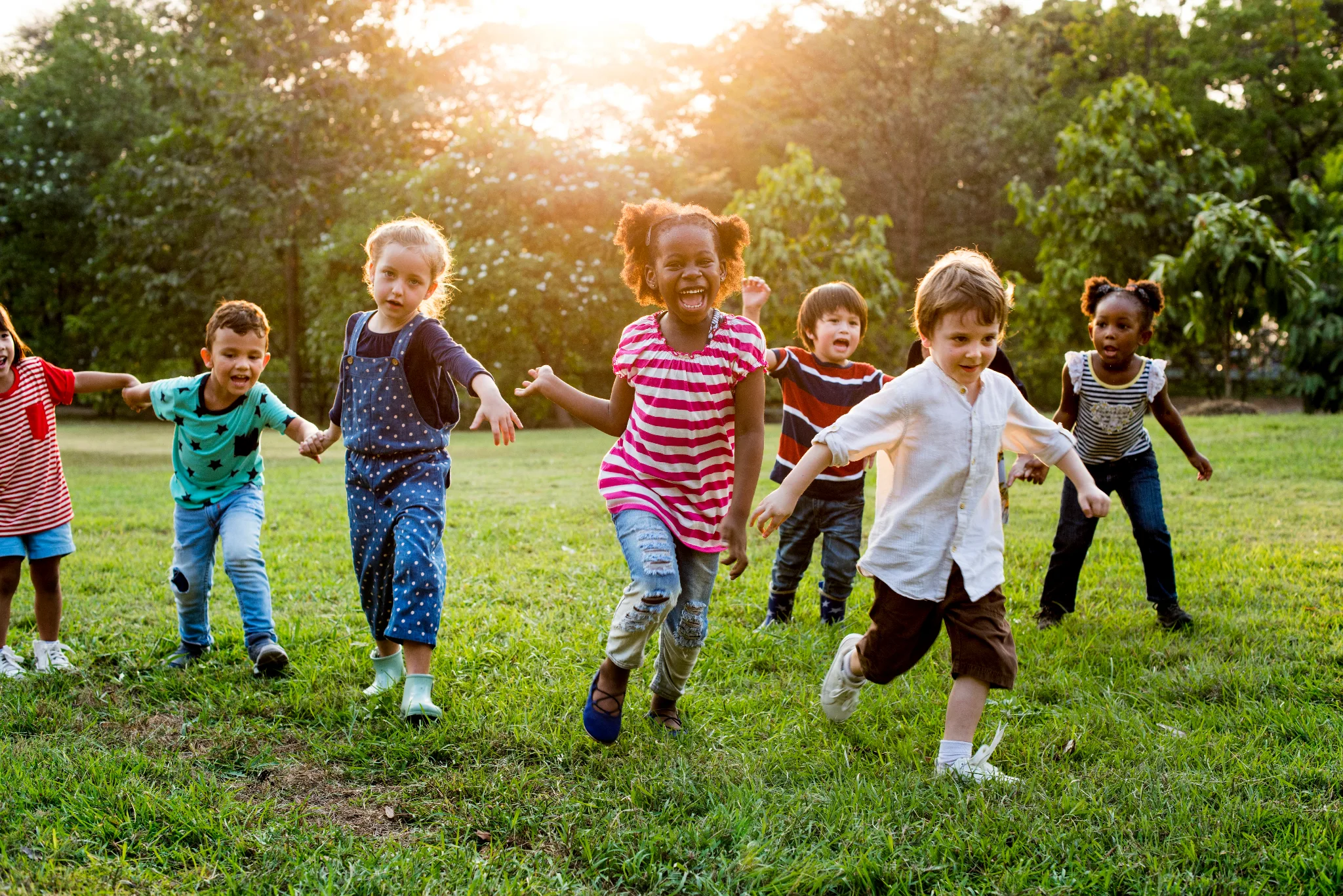 A diverse group of joyful young children running together across a sunlit grassy field with trees in the background.