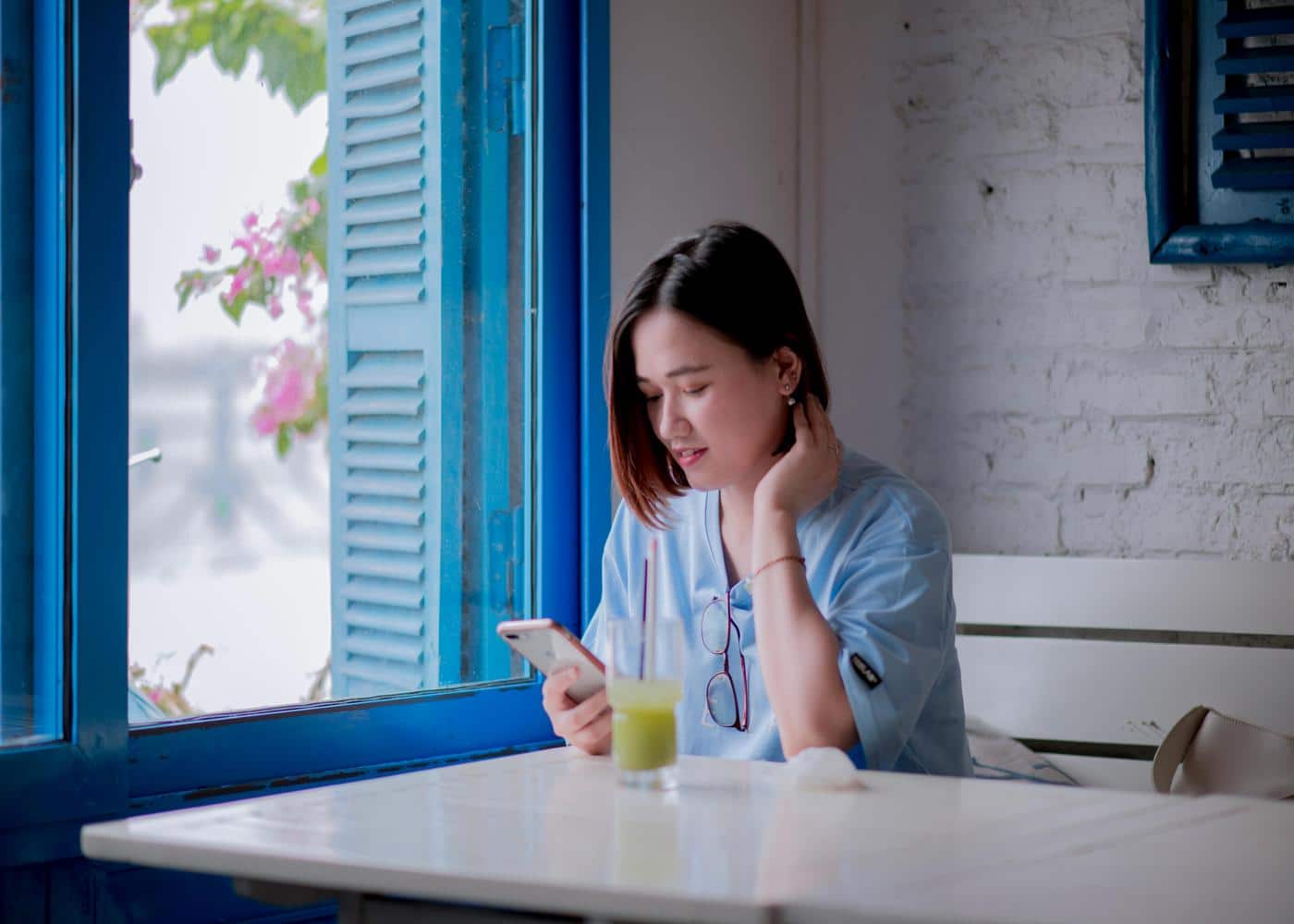Smiling woman sitting on white table next to blue window with a green juice looking at her phone