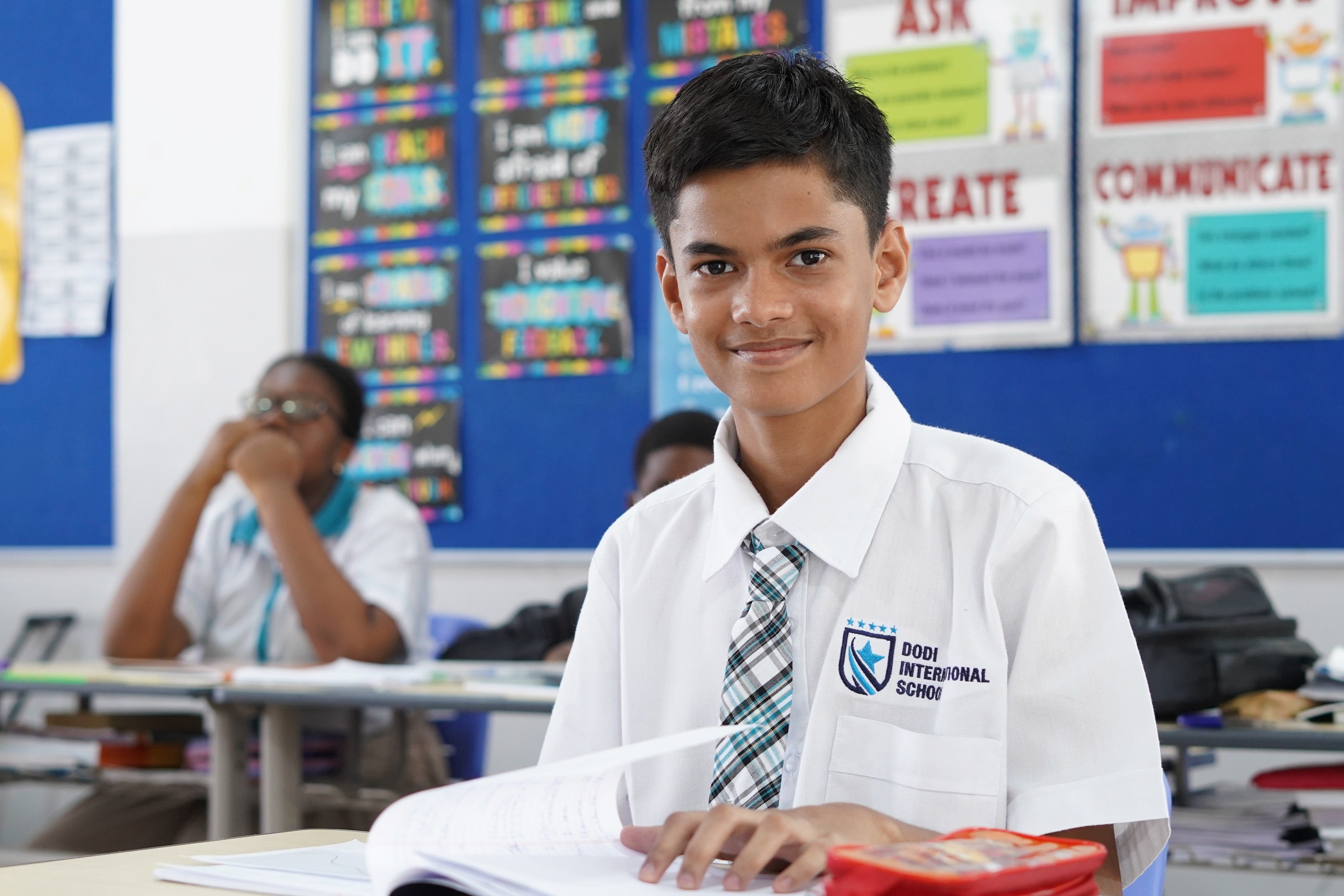 A smiling student in a white shirt and tie sits at a desk in a colorful classroom, engaged in learning.