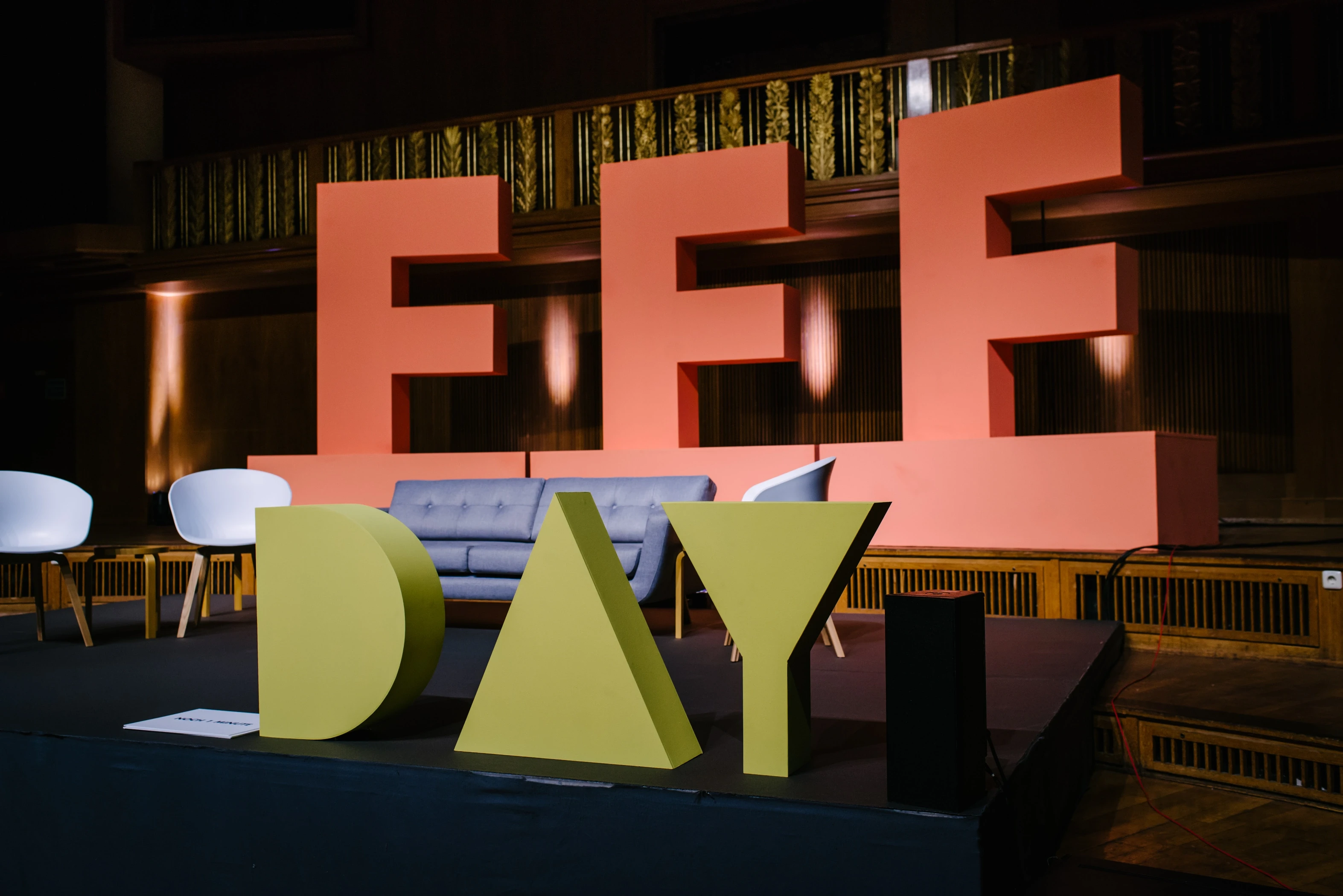 Close view of the stage set at FEMALE FUTURE FORCE DAY with giant coral FFF and olive-green DAY letters around a blue sofa and white chairs.