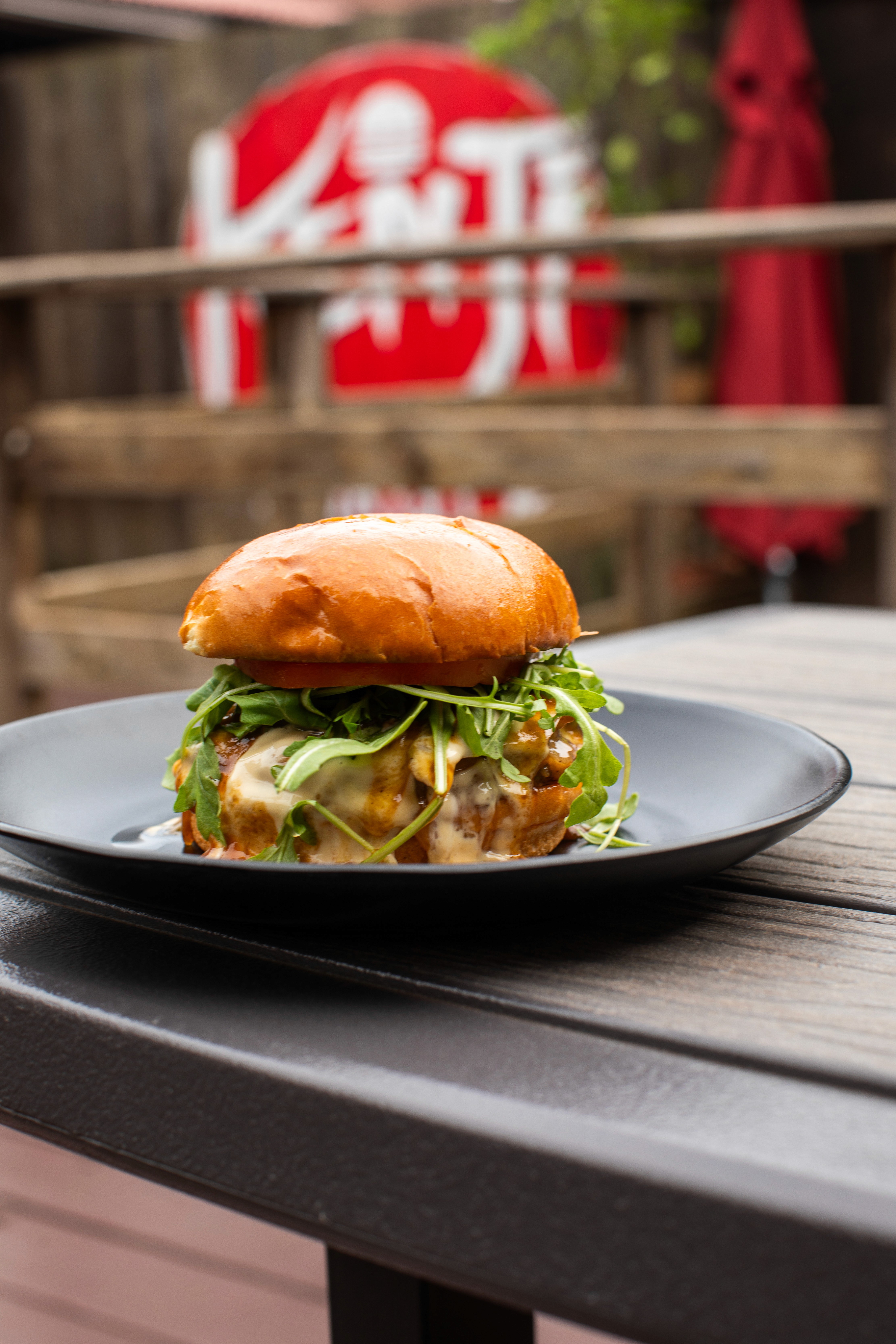 A rustic outdoor table setting featuring a charcuterie arrangement of cheese and sliced salami alongside a large bowl of uncooked pasta, a bottle of olive oil, and a glass of white wine, all against a dark, vine-covered background.