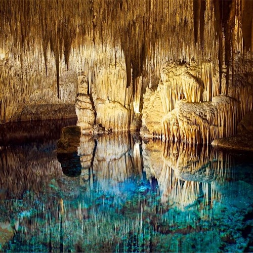 Una cueva con estalactitas y estalagmitas iluminadas reflejadas en una piscina subterránea clara de color azul.