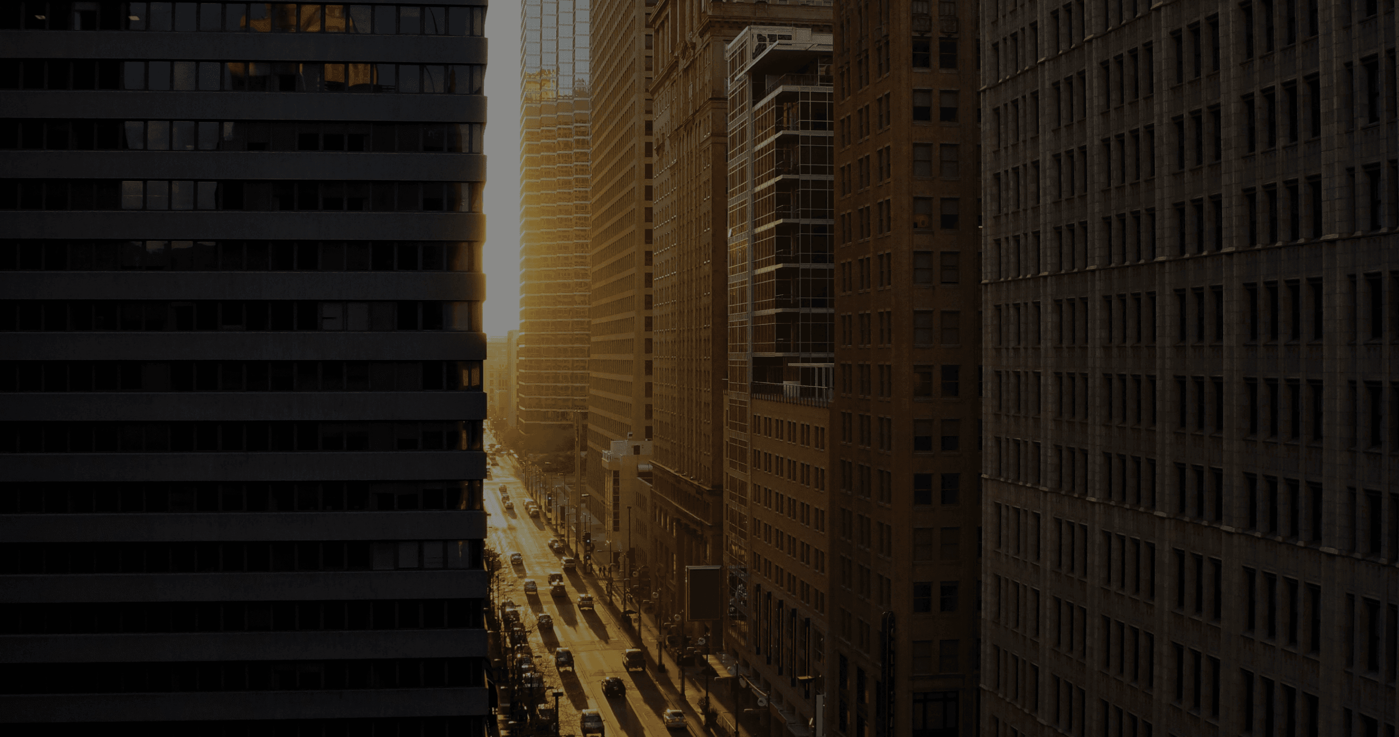 Aerial view of a city street lined with tall skyscrapers during sunset, with golden sunlight reflecting off the buildings and cars driving on the road below
