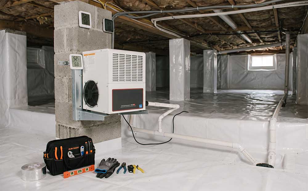A professional crawl space encapsulation and dehumidifier installation underneath a home. A white commercial-grade dehumidifier is securely mounted to a concrete block support pillar. The entire crawl space floor, walls, and pillars are completely sealed in a clean, bright white plastic vapor barrier liner. A white PVC drainage pipe connects to the unit and runs along the covered floor. In the foreground, an open black technician tool bag sits on the plastic liner alongside various hand tools, work gloves, and a roll of tape. The low ceiling shows exposed wooden joists and subflooring.
