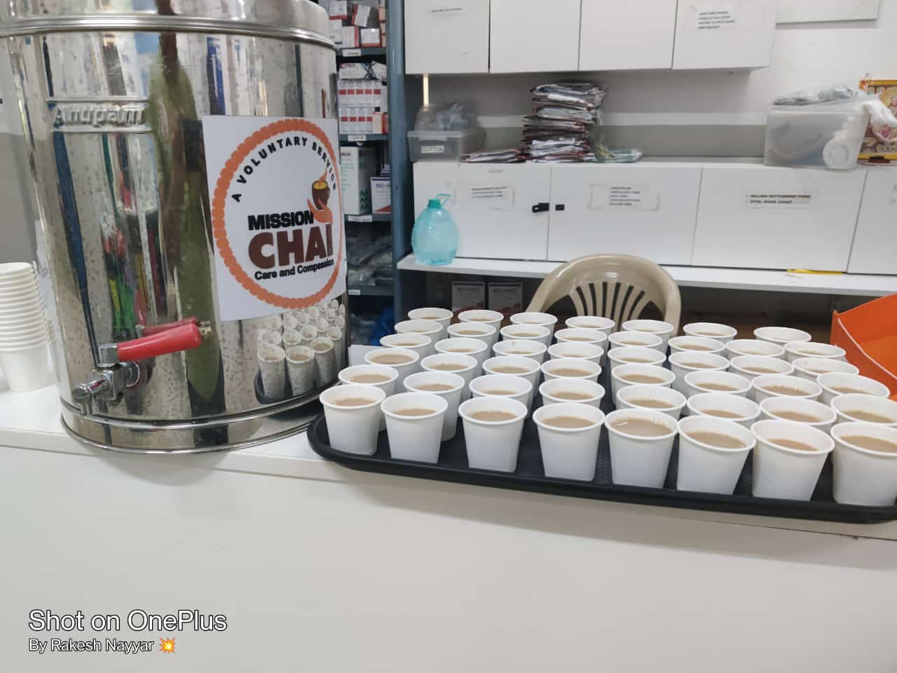 Large metal dispenser labeled "Mission Chai" next to a tray of small paper cups in a hospital corridor.