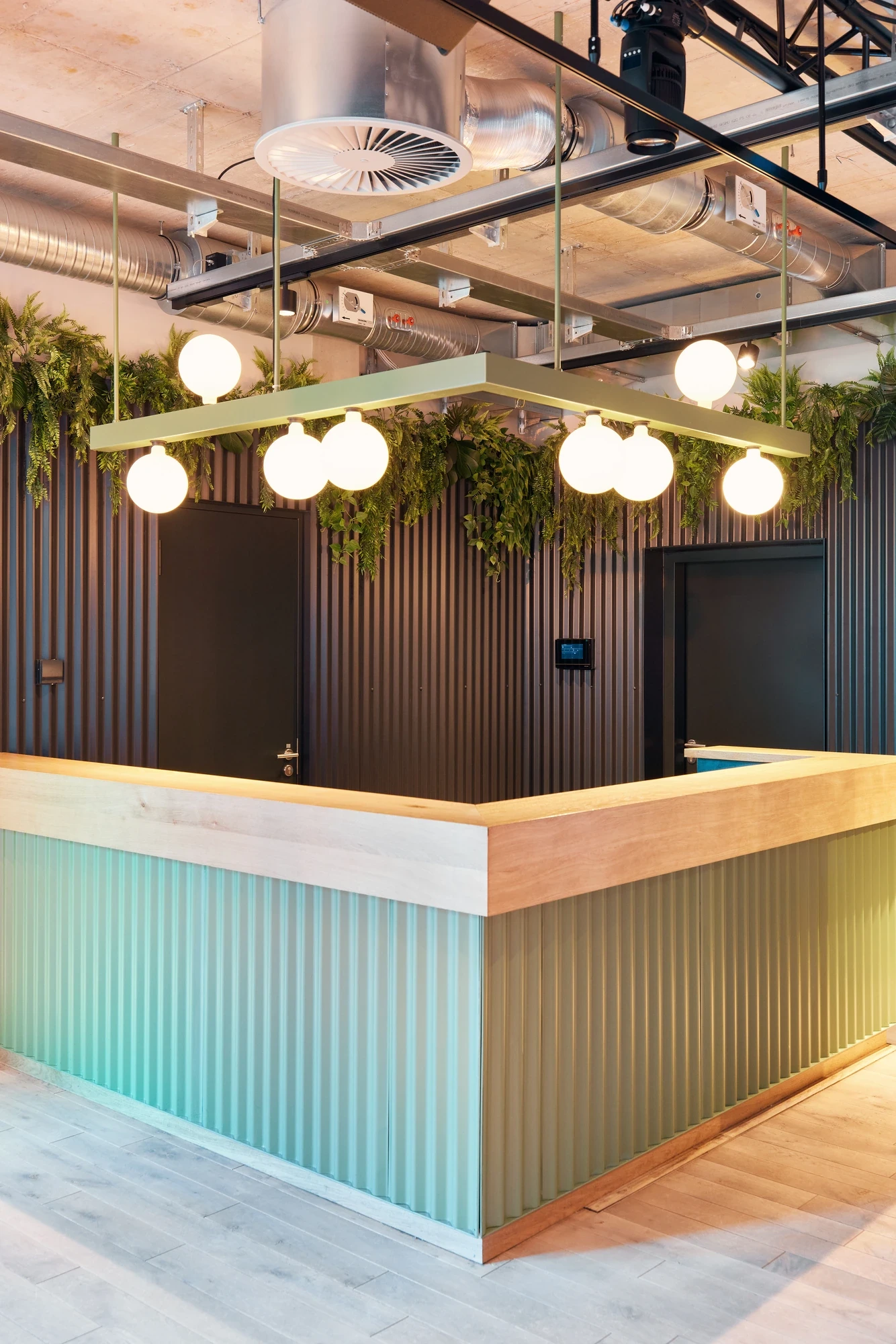 Event counter with mint-green corrugated cladding and wooden top, framed by a rectangular pendant fixture with globe lights and trailing greenery.