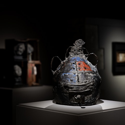 Isang makinis na helmet na gawa sa madilim na metal na may pulang at asul na disenyo na nakadisplay sa pedestal sa isang madilim na ilaw na eksibit.