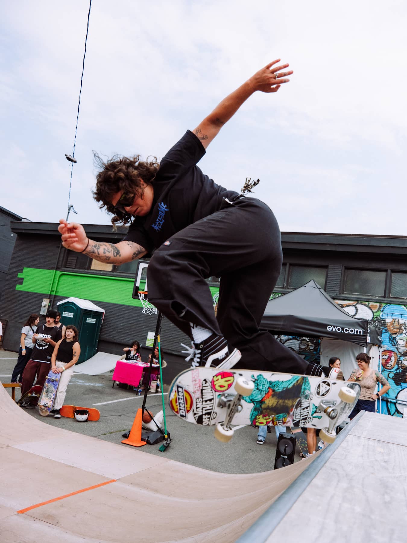 A skateboarder in black clothing and sunglasses performs a trick on a ramp at an outdoor skatepark with spectators in the background.
