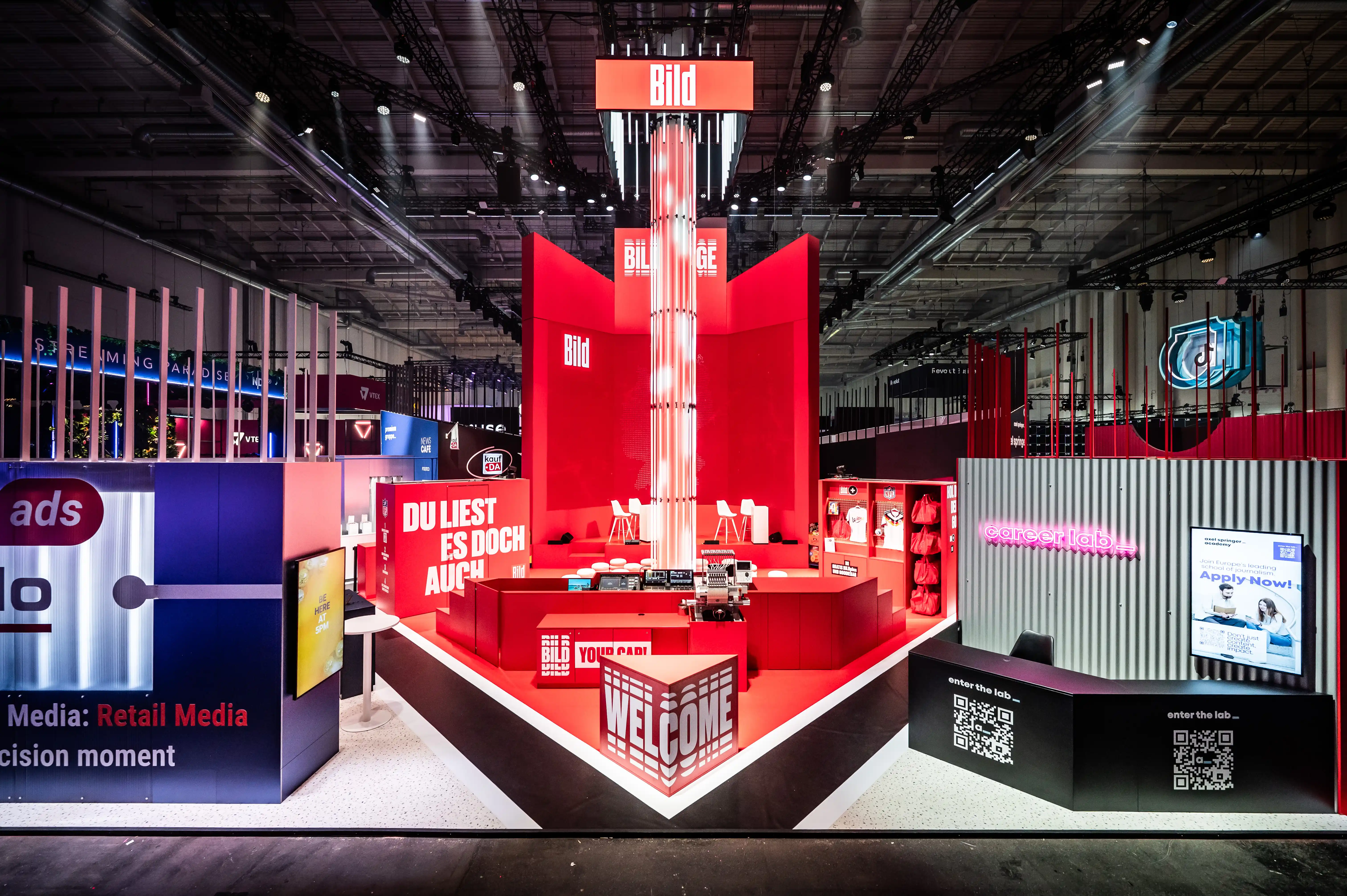 Front view of the red Bild booth with illuminated central tower, Welcome sign and surrounding partner areas.