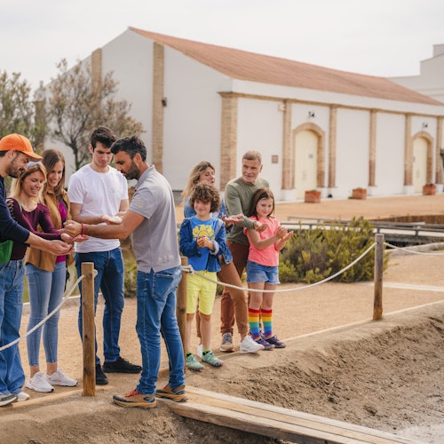 Eine Gruppe von Erwachsenen und Kindern beobachtet einen Mann, der etwas in der Nähe einer kleinen Brücke in einer historisch anmutenden Umgebung erklärt.