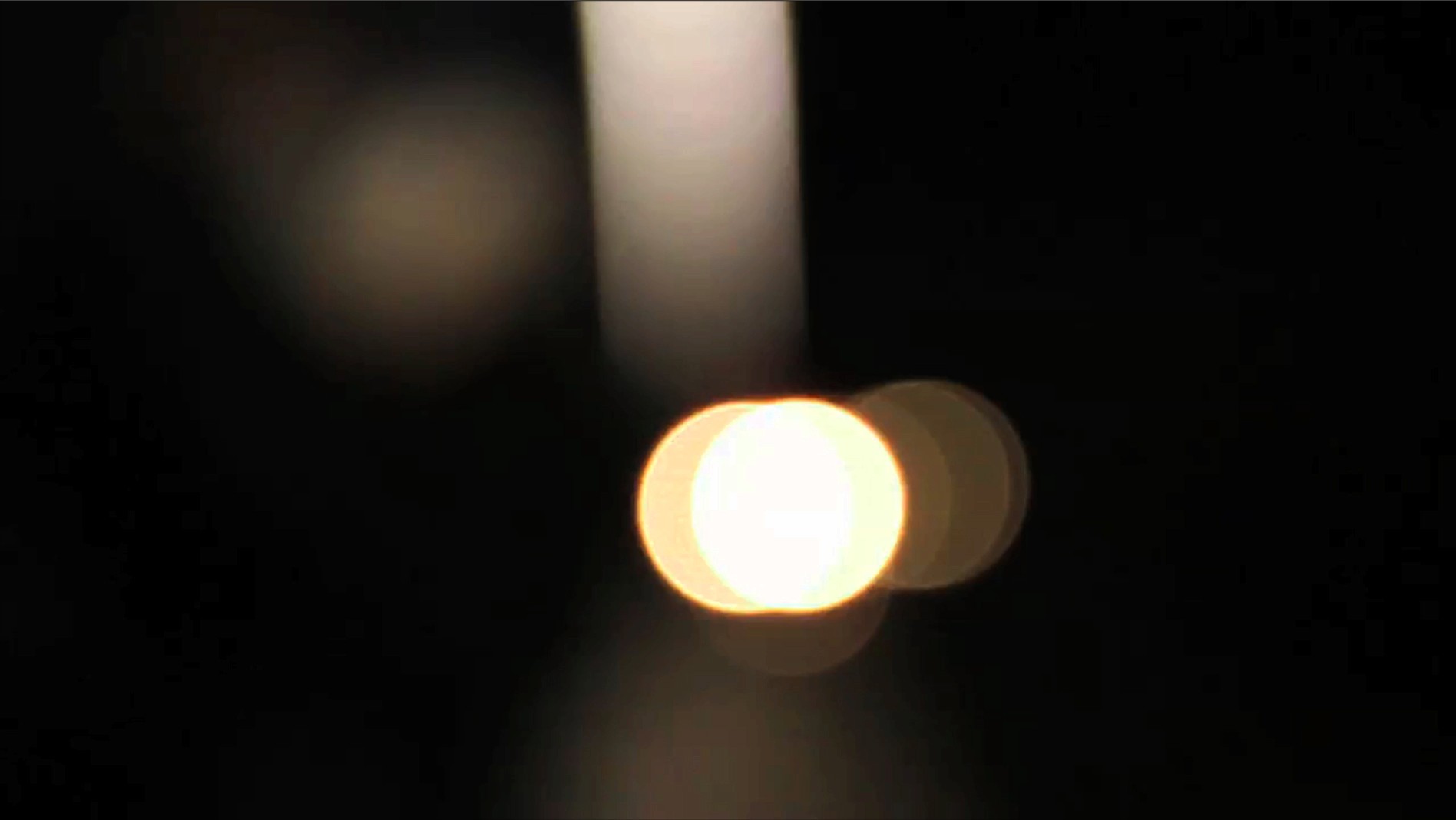 A bright, circular light source emits a warm glow against a dark background. The light appears slightly blurred or out of focus, with concentric rings visible around its perimeter. The background is predominantly black, with some faint, diffused light areas visible to the left of the central light source.
