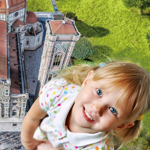 A smiling young girl with blonde hair appears giant next to a detailed miniature model of a historical building and lush greenery.