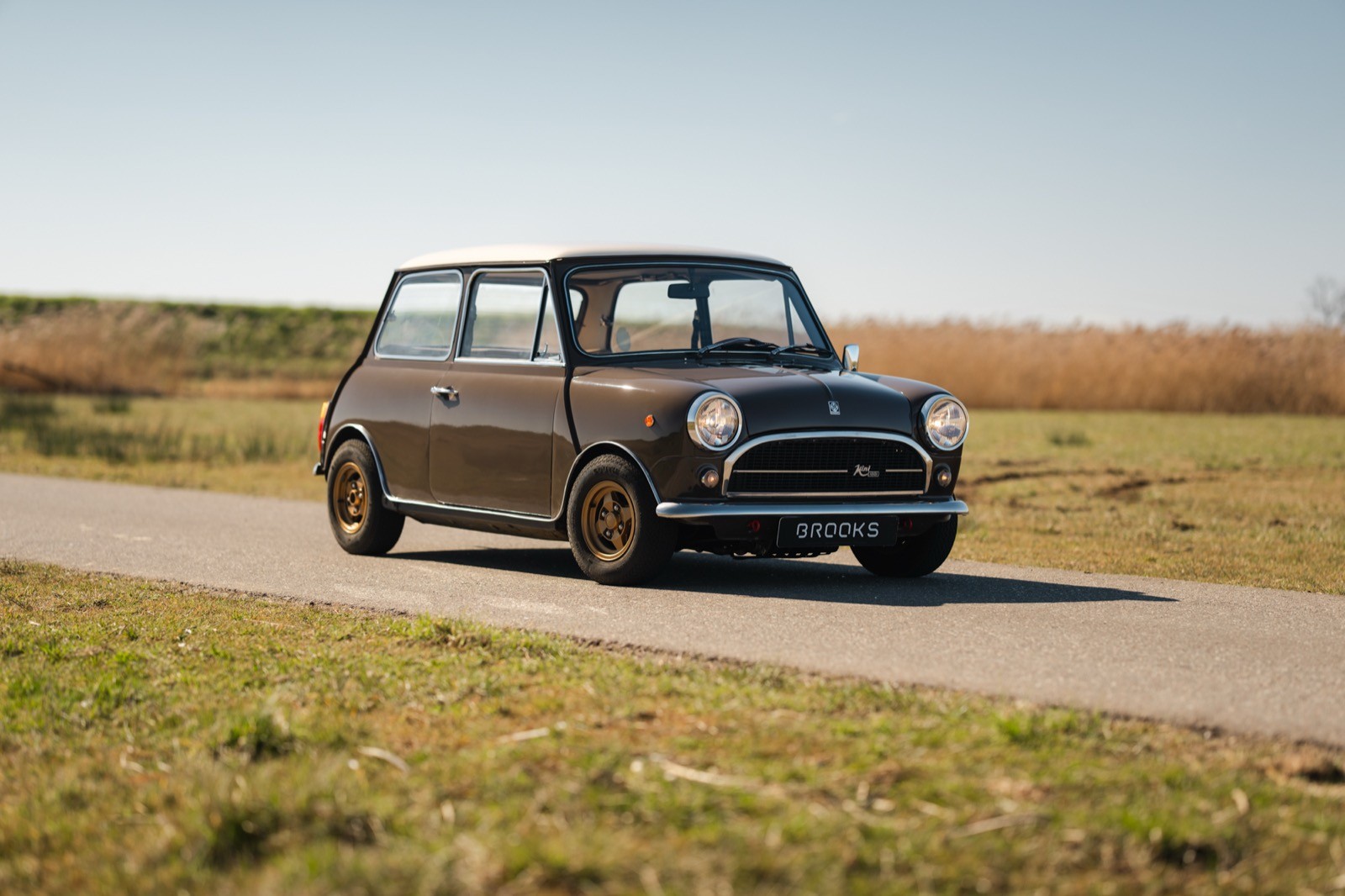 A vintage car parked on a rural road, surrounded by open fields and a clear blue sky.