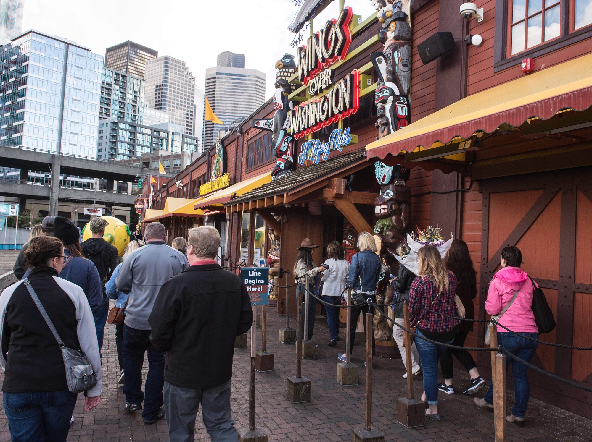 Visitors queue under the neon Wings Over Washington sign and carved totem poles on the Pier 57 boardwalk.