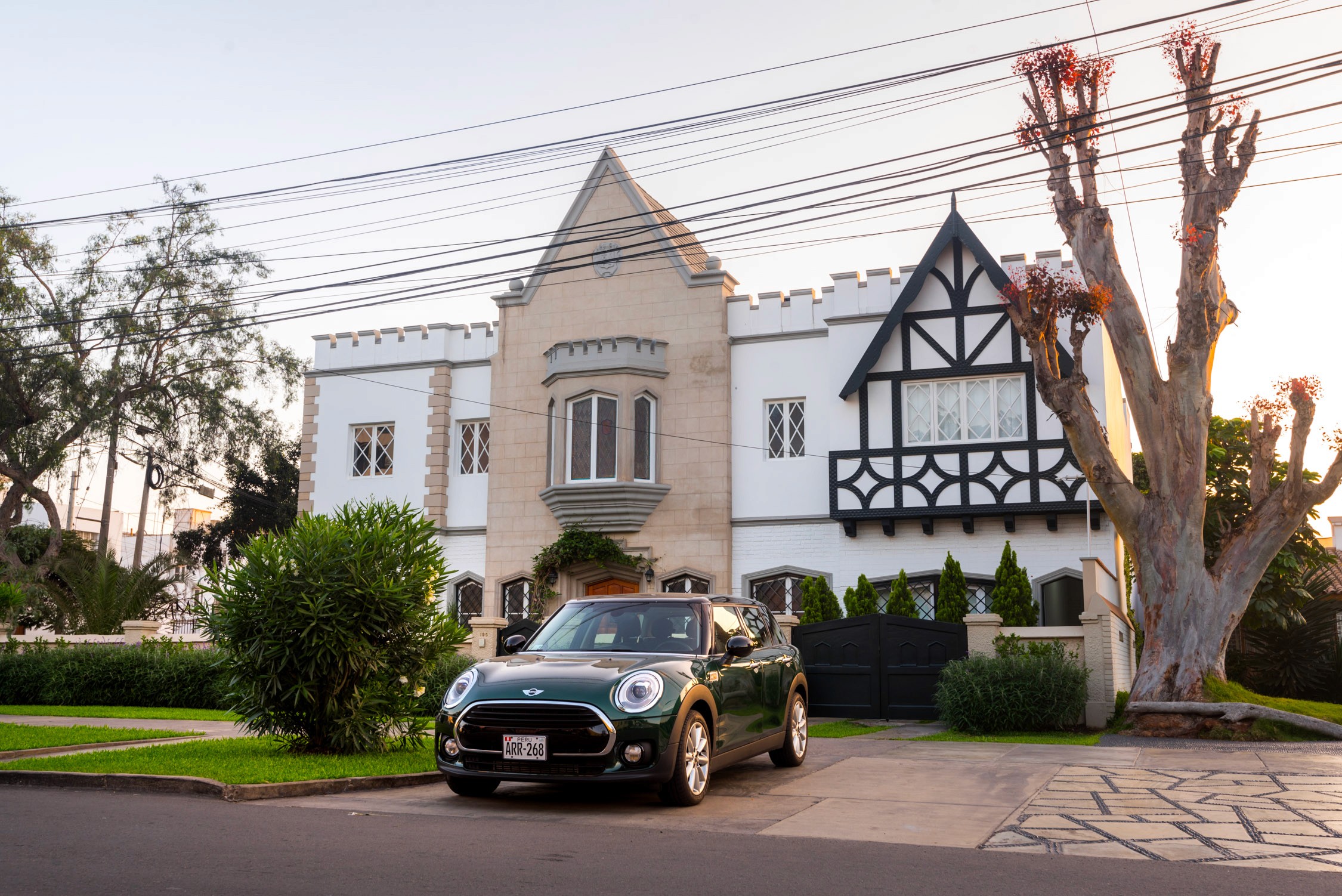 A green Mini Cooper parked in front of a large white and tan Tudor-style mansion with a gabled roof and arched windows.