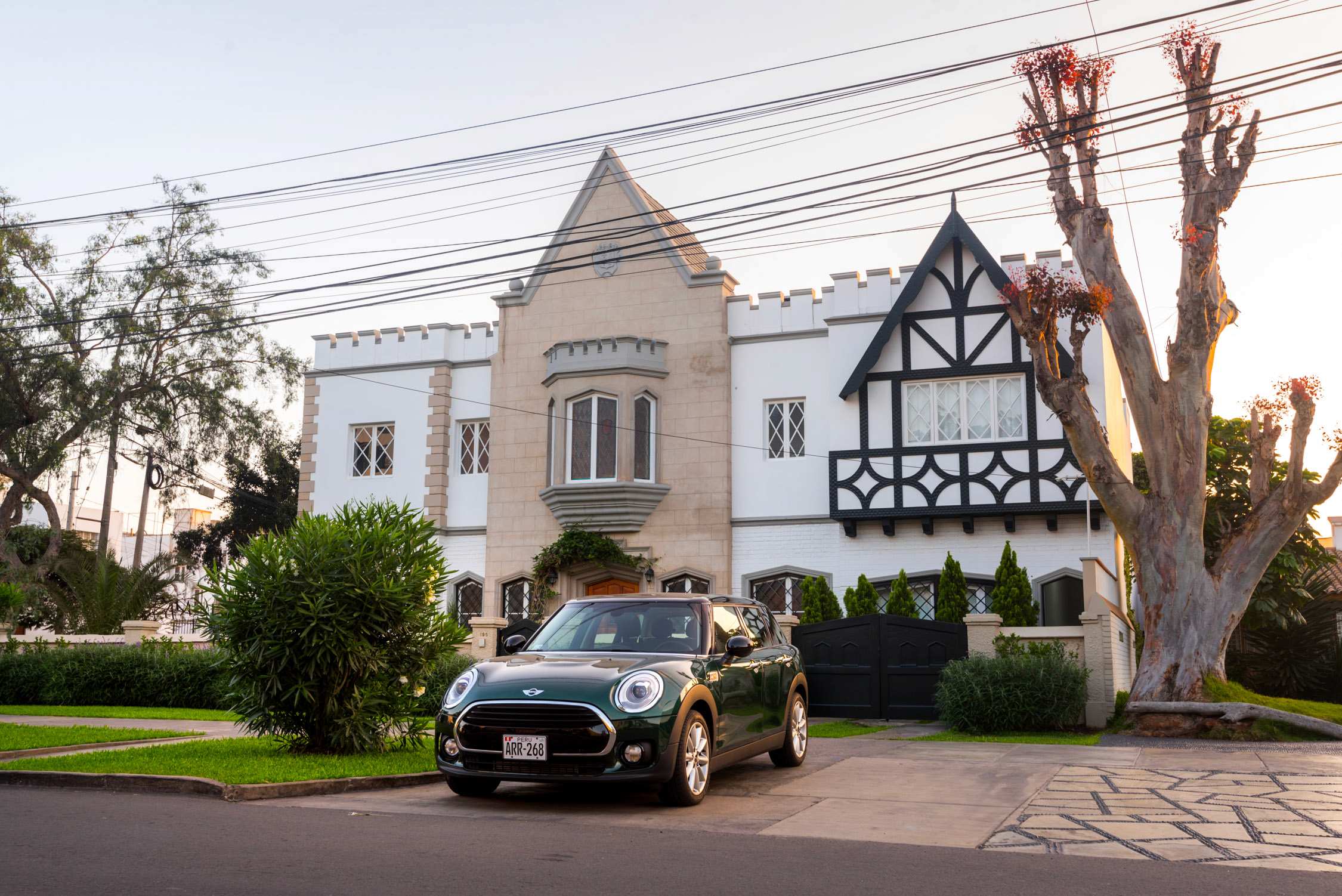 A green Mini Cooper parked in front of a large white and tan Tudor-style mansion with a gabled roof and arched windows.