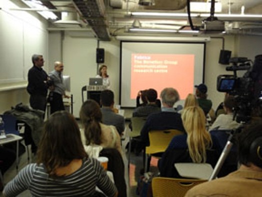 A photo of a group of people listening to a lecture and watching a screen that has an orange projection on it with the white text.