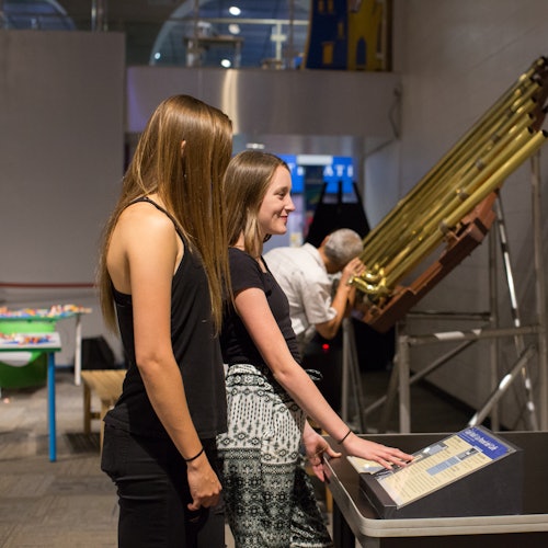 Dos mujeres jóvenes interactúan con una exhibición que presenta tubos de latón y un panel de control. Un hombre mayor observa al fondo.