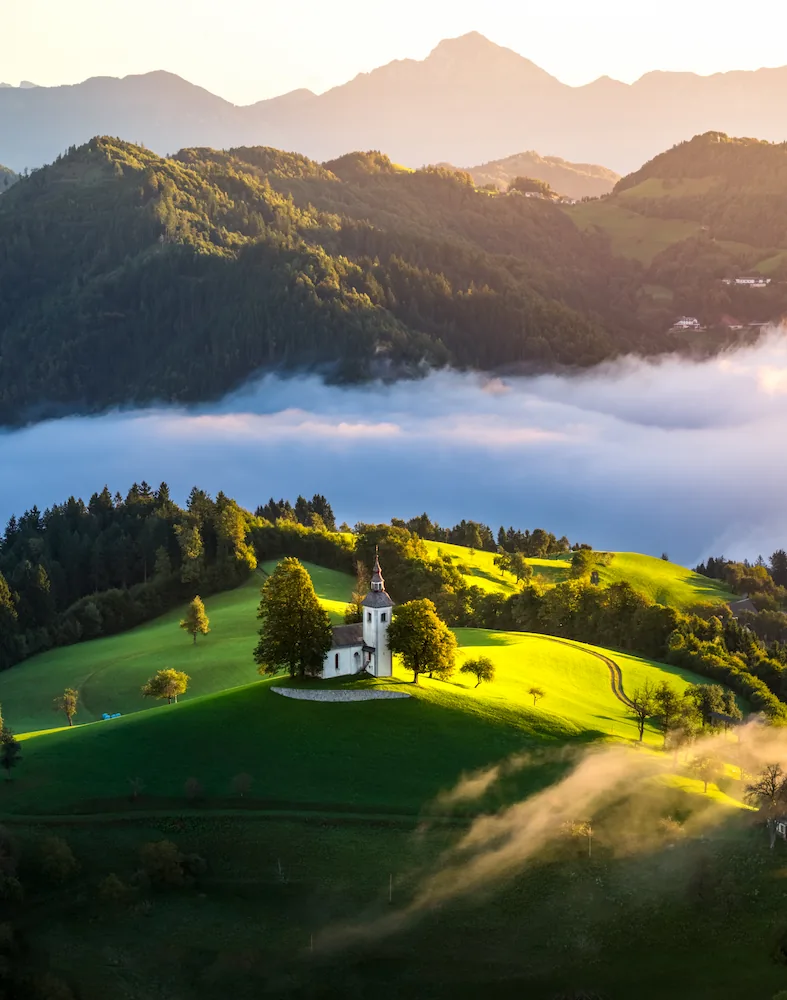 Aerial view of the Church of Saint Thomas (Sveti Tomaž) on a lush green hilltop in Slovenia at sunrise, with morning mist in the valleys and the Kamnik-Savinja Alps glowing in the background.