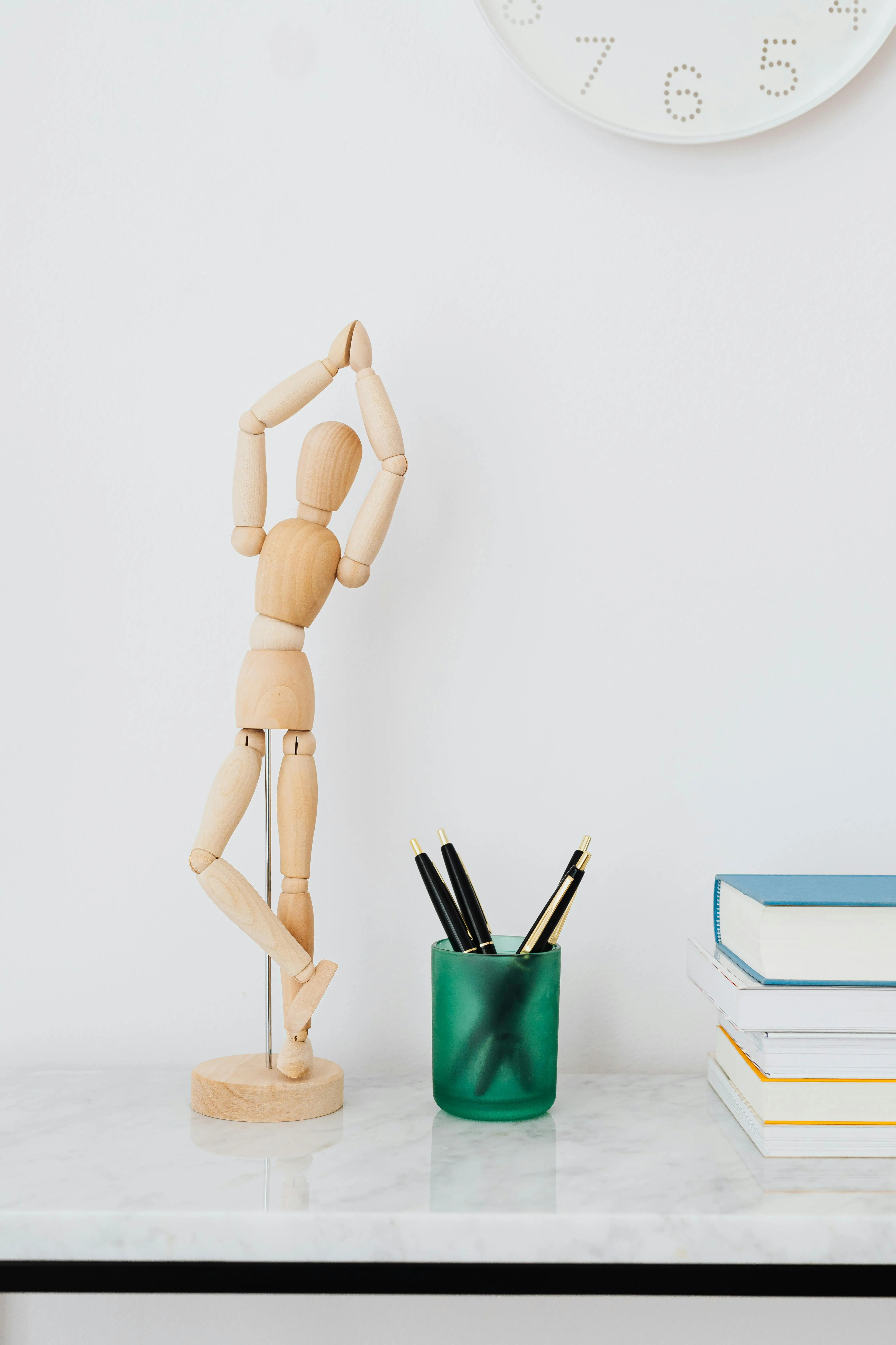 A wooden artist's mannequin stands on a desk next to a green cup of pens and a stack of books.