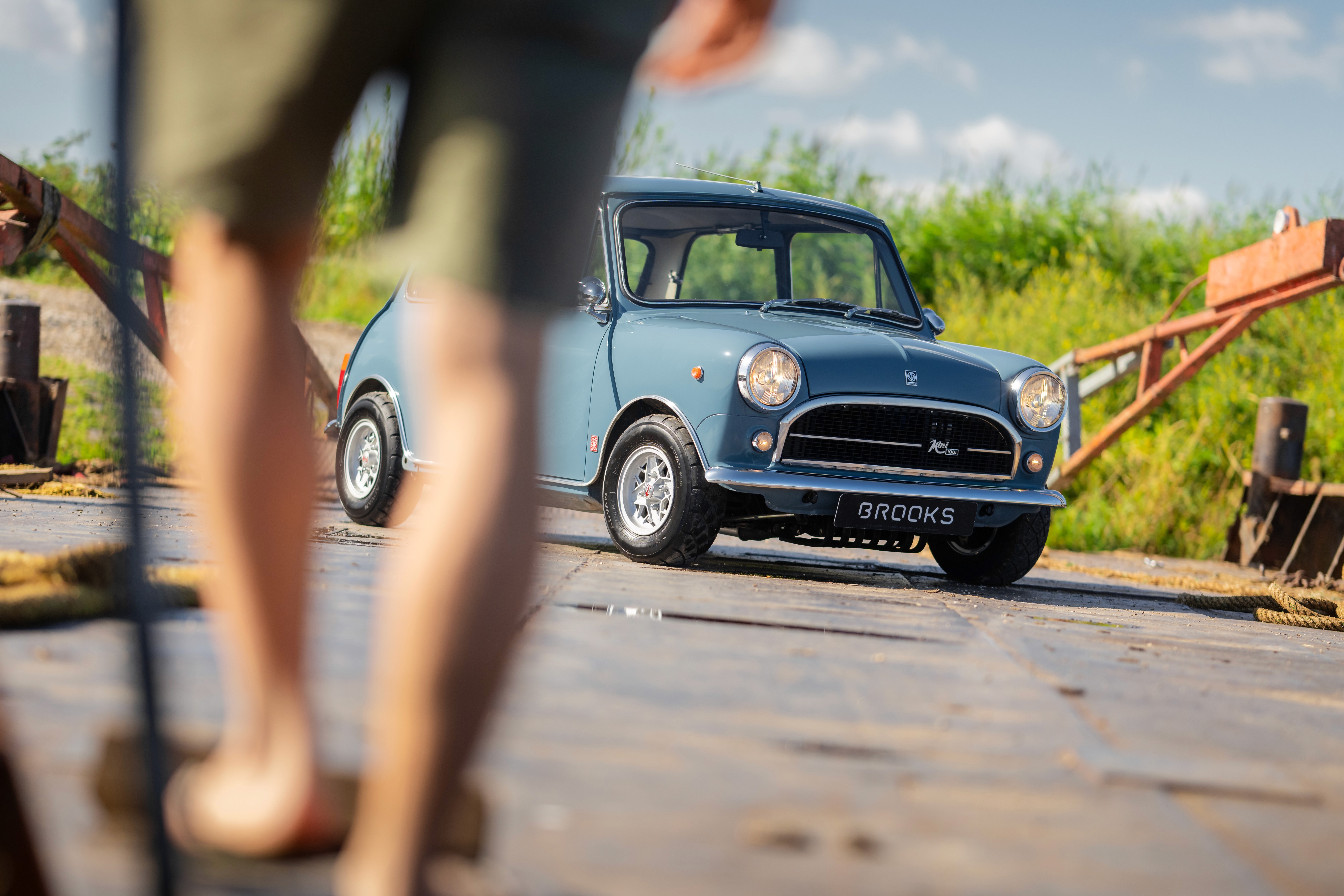 A person in shorts walks toward a blue vintage car parked on a sunny day with greenery in the background.