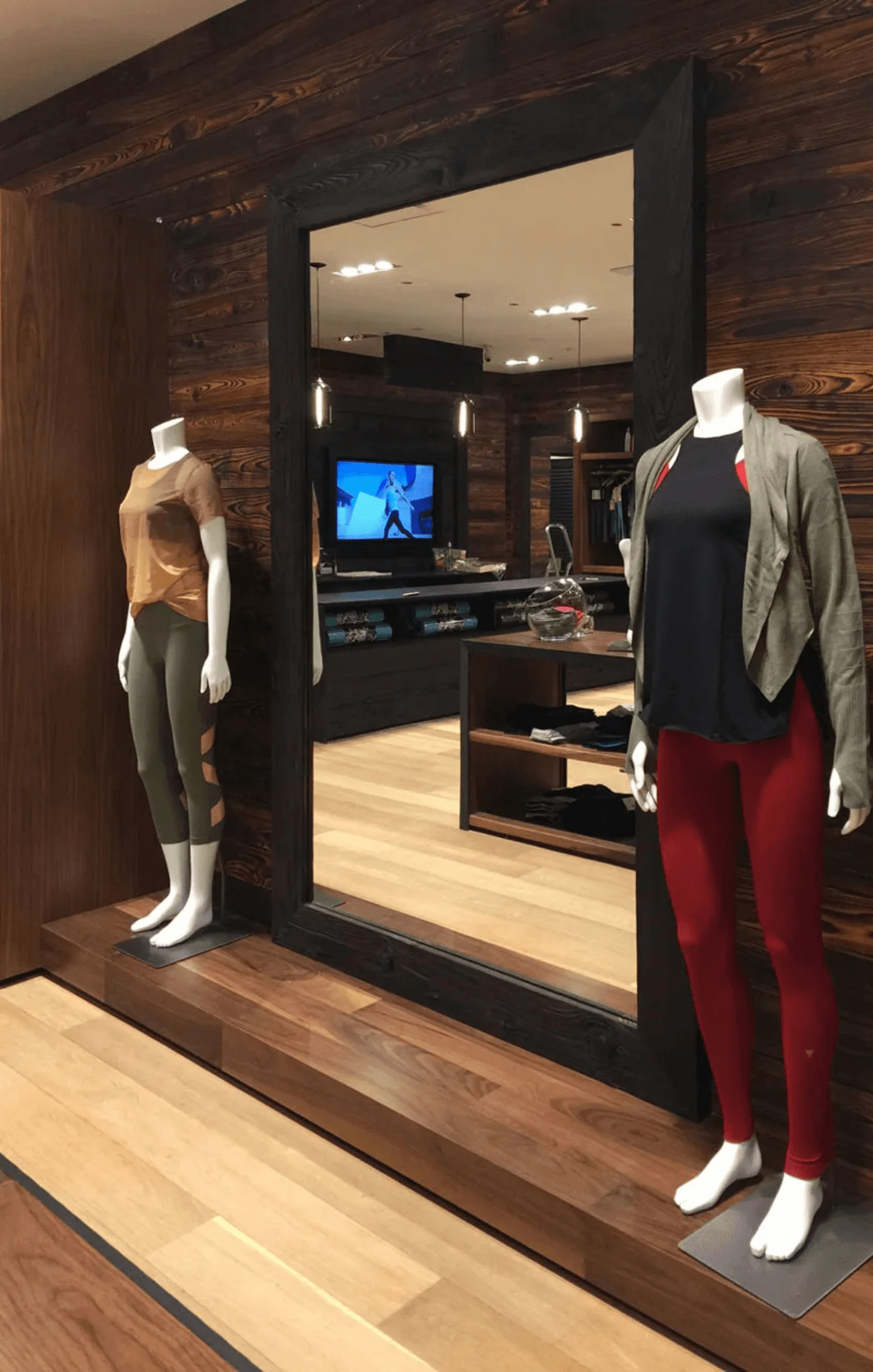 Mannequins in YOGASMOGA activewear stand before a large mirror framed in dark wood, with a video display reflected behind