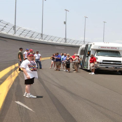 Grupper af mennesker står på en racerbane ved siden af en shuttlebus med teksten "World Fan Tours" skrevet på.