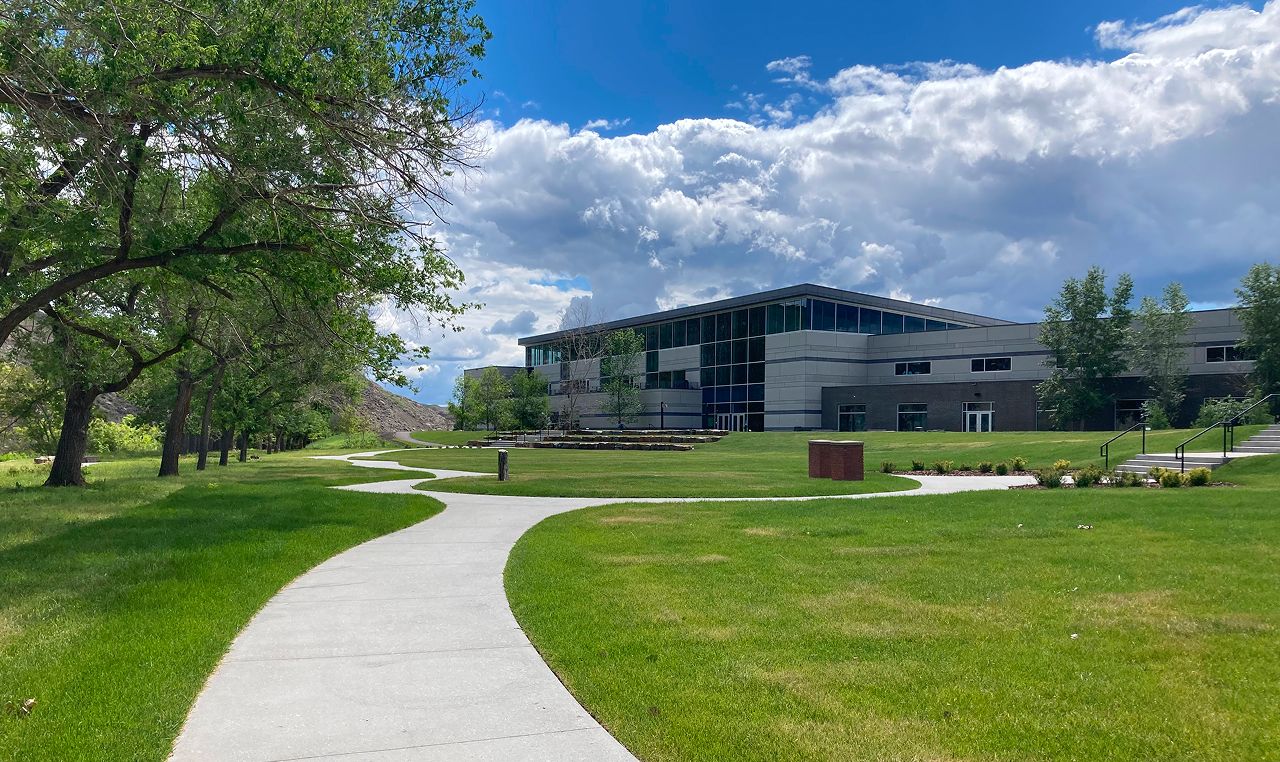 Completed multi-use pathway system and landscaping at Drumheller Downtown Dike project with modern building and mature trees under dramatic prairie sky