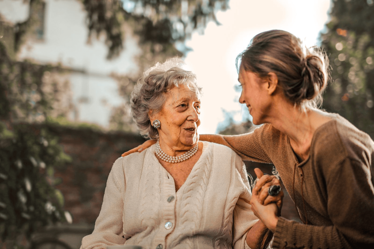 An older woman and her daughter laugh together in the garden, a hand resting on her shoulder.