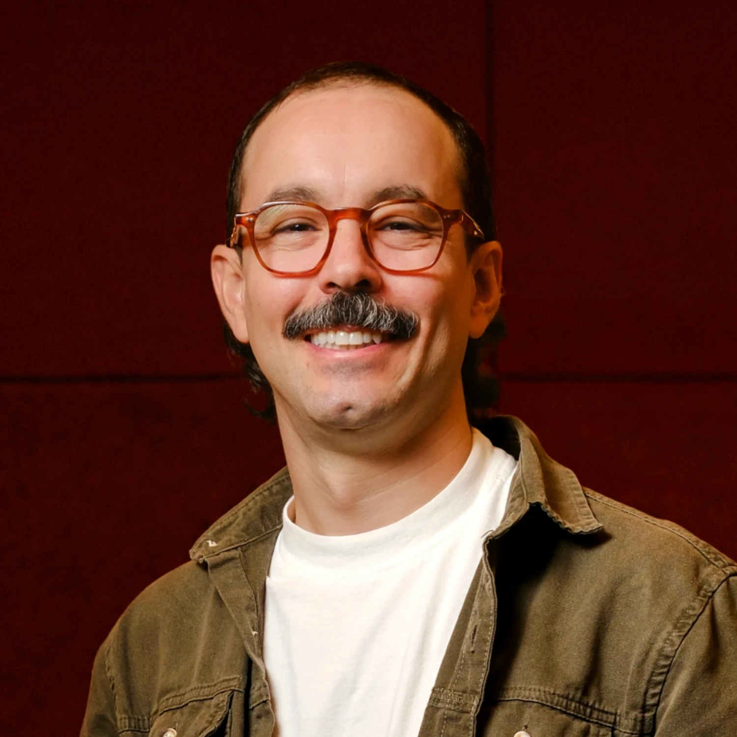 A young person with curly hair smiles at the camera while sitting in front of a laptop against a dark background.