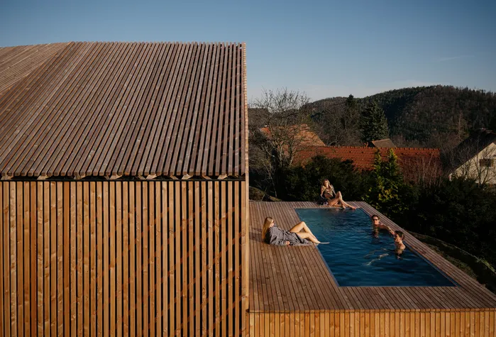Hoam Chalet mit Lärchenfassade, davor Gäste am Pool auf der Holzterrasse mit Blick auf die Hügel.