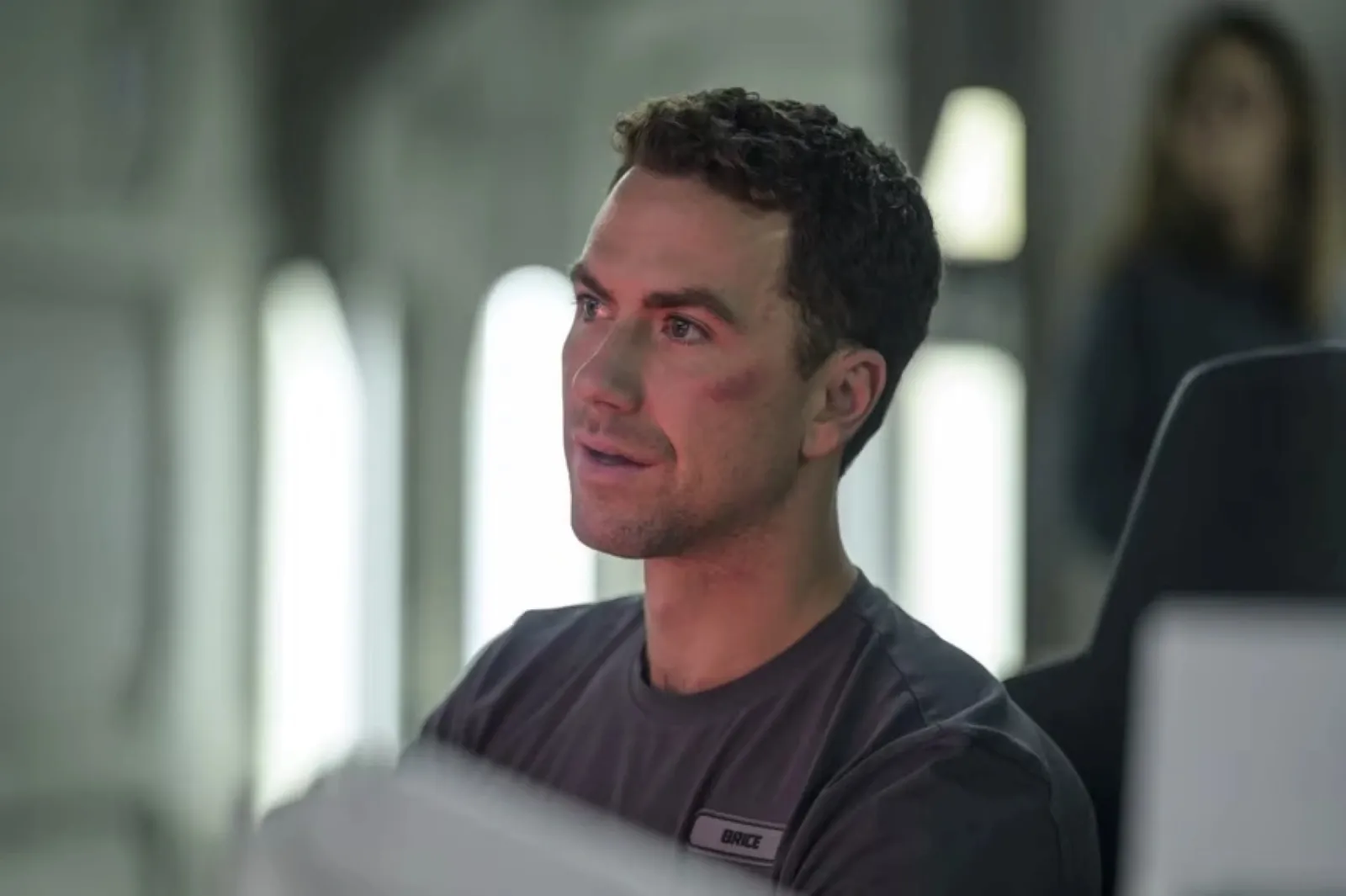 A man with short, dark hair is sitting in a dimly lit control room setting, looking thoughtfully towards the side, wearing a dark-coloured shirt, with blurred lights and furniture in the background.