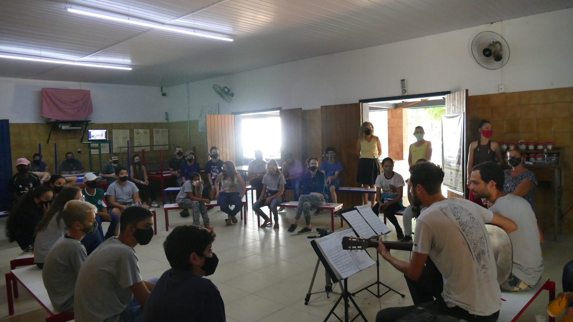 Músicos com violão e partituras se apresentam para alunos de máscara em salão de escola