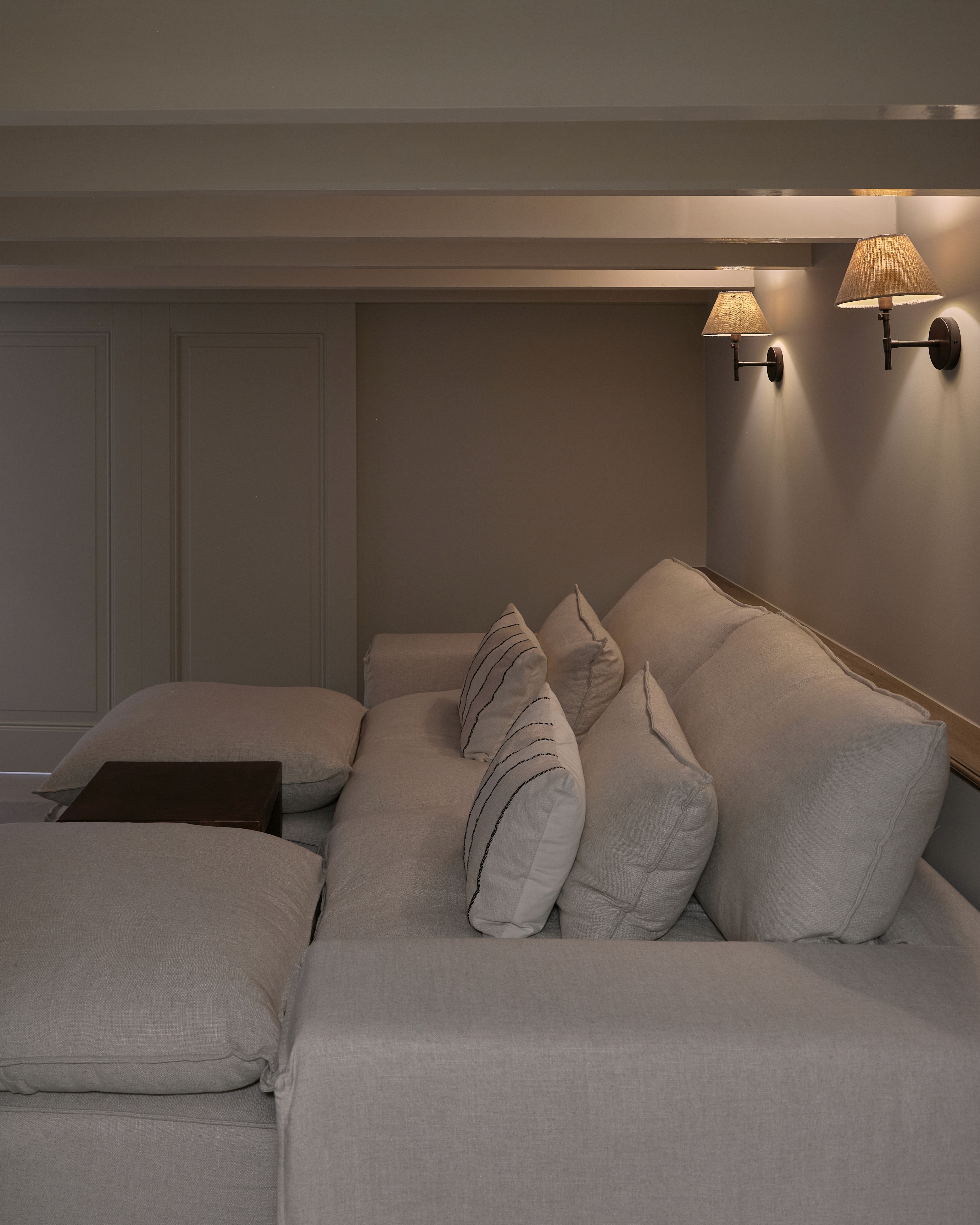 Cosy cinema seating area with linen sectional sofa, layered cushions, wall sconces with natural linen shades, and exposed ceiling beams