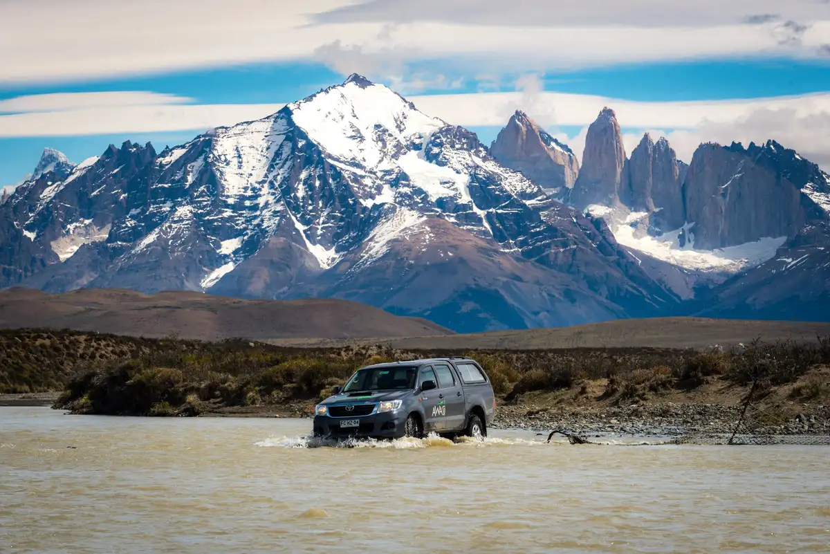 A silver SUV drives through a shallow river in front of snow-capped mountains and jagged granite peaks under a blue sky.