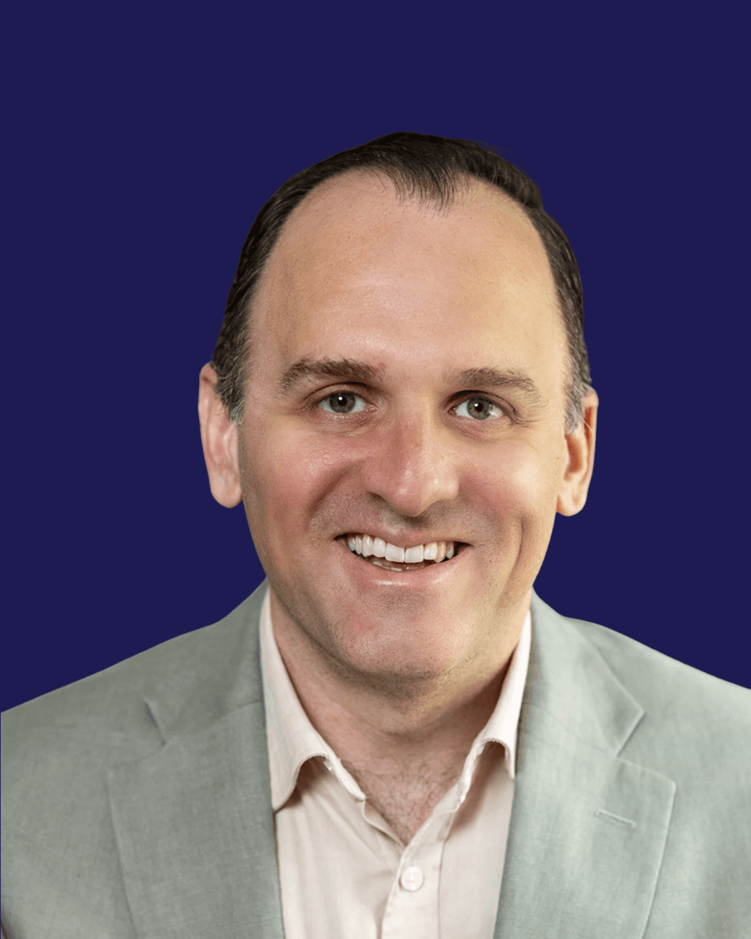 A smiling person with short dark hair wears a light gray blazer and a beige shirt, set against a solid dark blue background.