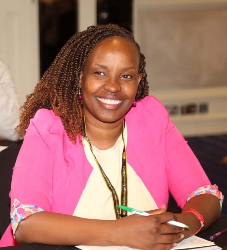 A smiling young woman with curly hair, wearing a purple shirt, stands in a bright indoor setting.