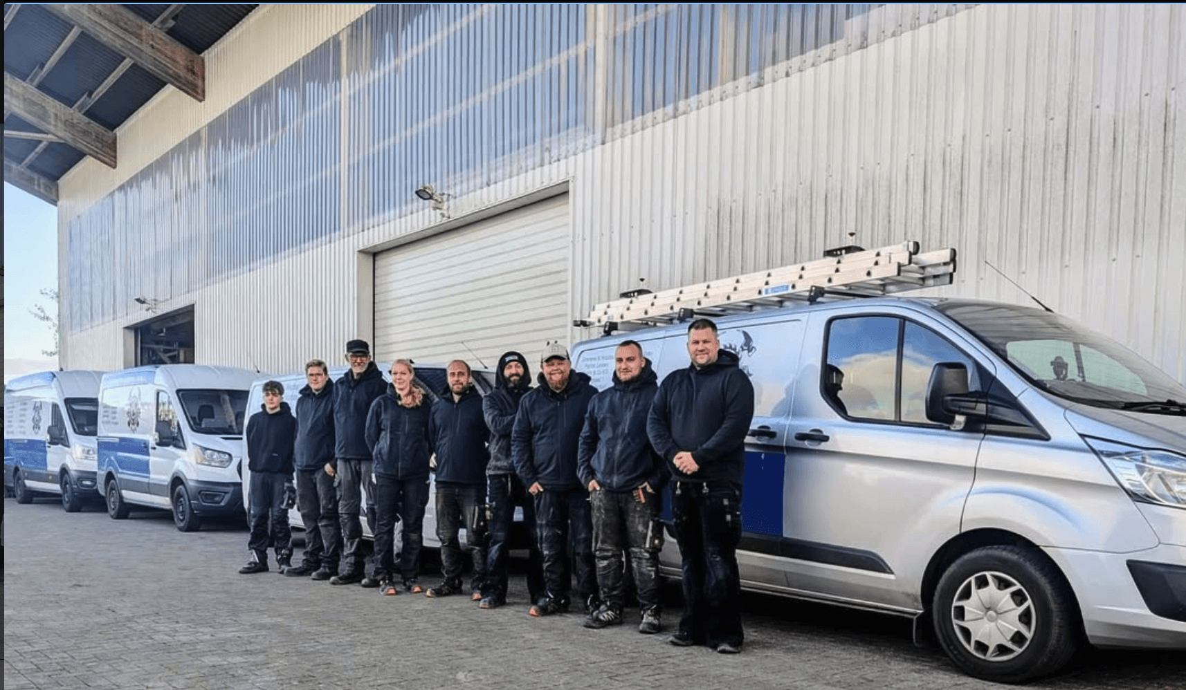 Eine Gruppe von Arbeitern in dunklen Uniformen steht in einer Reihe neben mehreren Firmenwagen, die mit Leitern ausgestattet sind und auf einem befestigten Untergrund vor einem großen Lagerhaus geparkt sind.