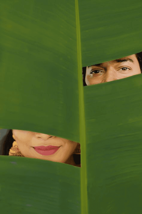 Creative contemporary wedding portrait of a couple peeking through green palm leaves, showcasing a playful and modern approach to wedding photography.