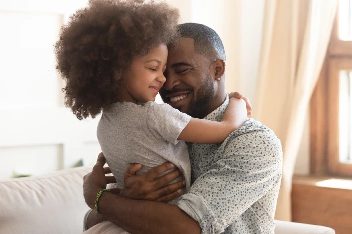 A imagem mostra um momento afetuoso entre um homem e uma criança, possivelmente pai e filha, enquanto eles se abraçam com sorrisos de felicidade. A menina, com cabelo cacheado volumoso, veste uma camiseta cinza e está de olhos fechados, aproveitando o carinho. O homem, usando uma camisa clara com padrões, também sorri com os olhos fechados, transmitindo uma sensação de amor e conexão. A iluminação suave e as cortinas claras ao fundo contribuem para criar um ambiente aconchegante e familiar, destacando o vínculo emocional entre os dois.