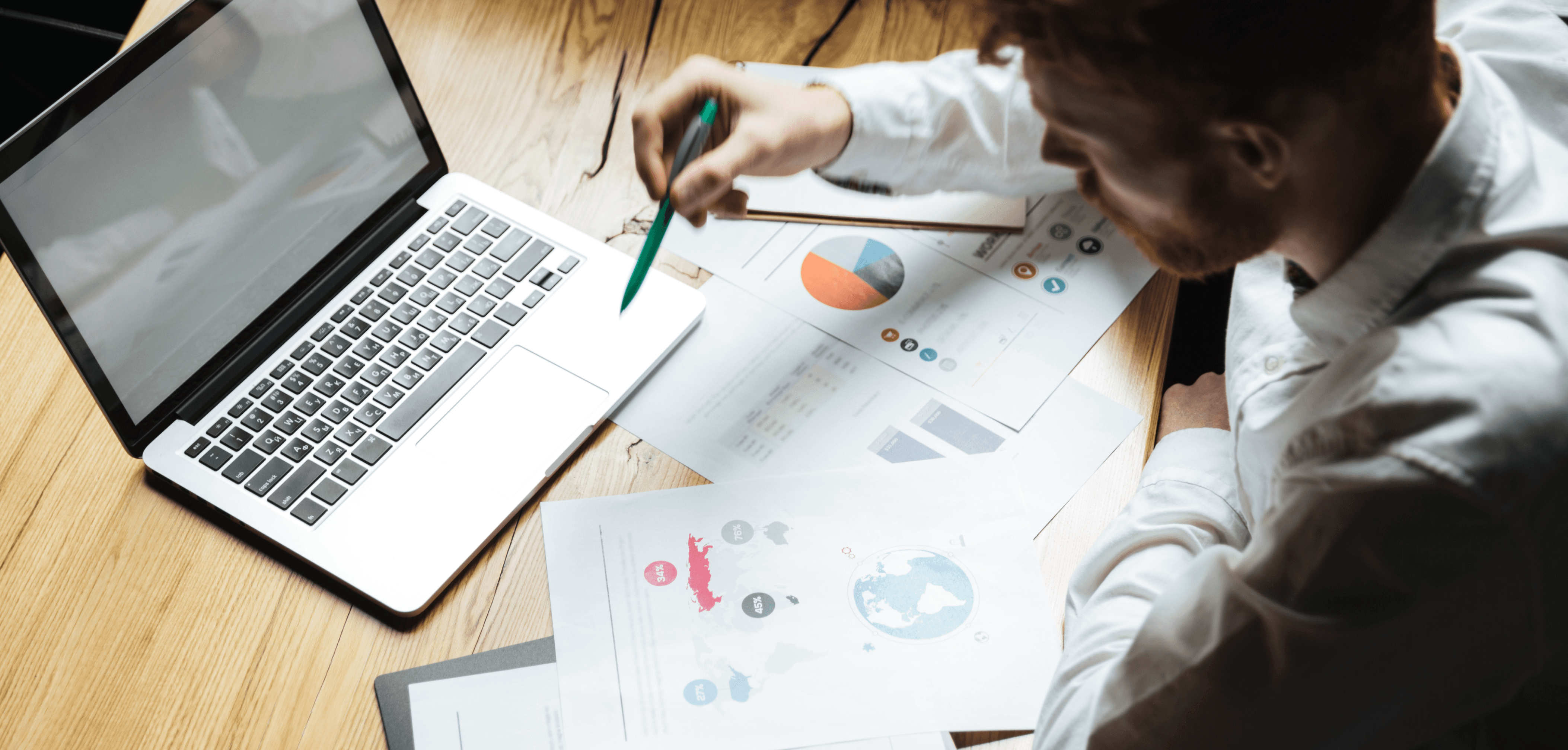Person reviewing printed charts and graphs beside an open laptop, pointing at data with a pen on a wooden desk.