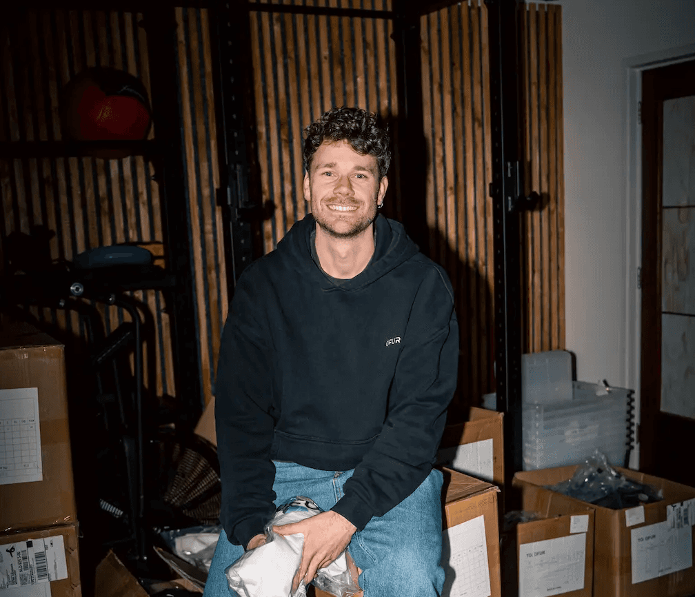 Co-founder van OFUR zit lachend in een zwarte hoodie tussen de verzenddozen in het magazijn
