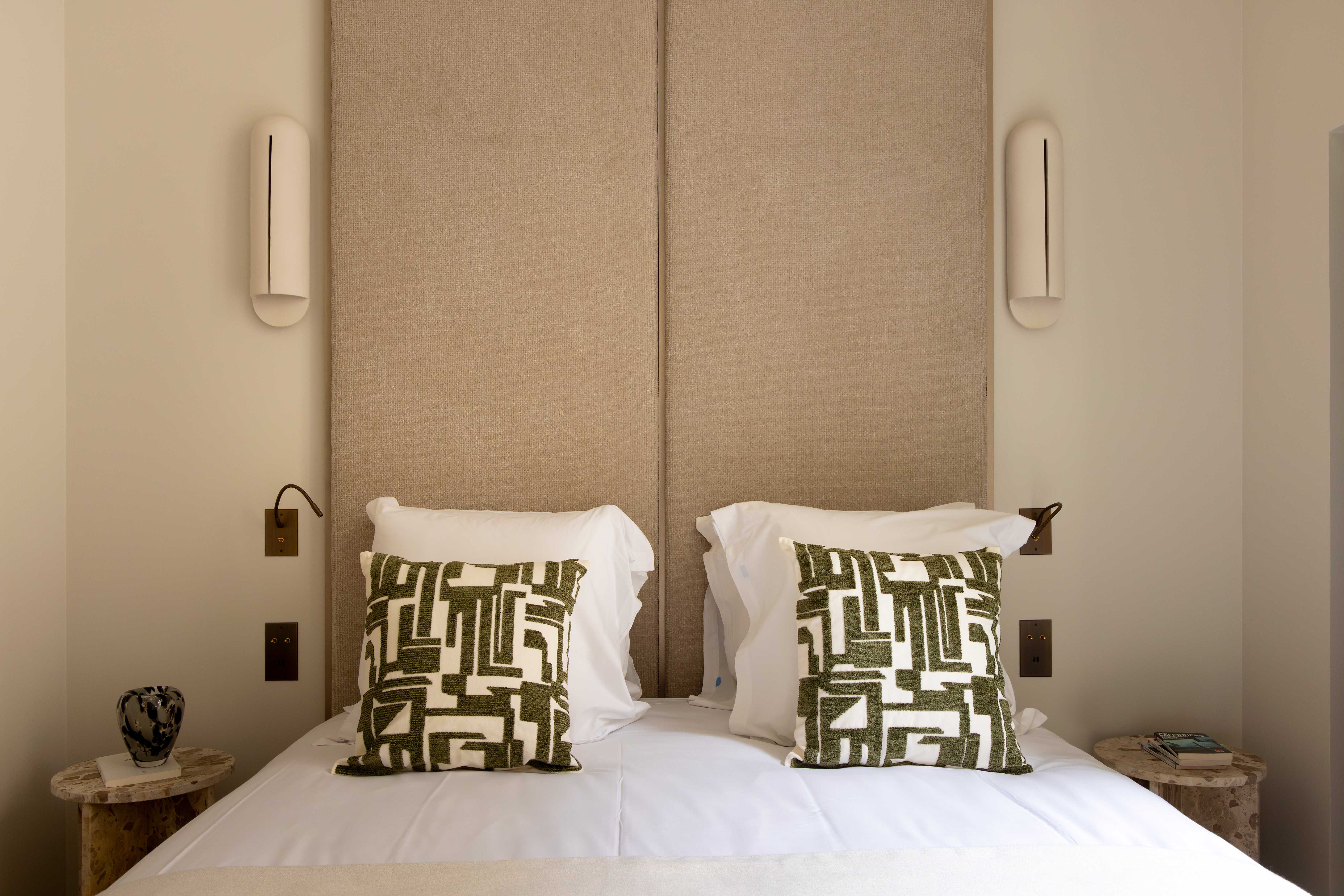 Detail de la tete de lit beige avec coussins graphiques verts, appliques ovales et tables de chevet en bois petrifie