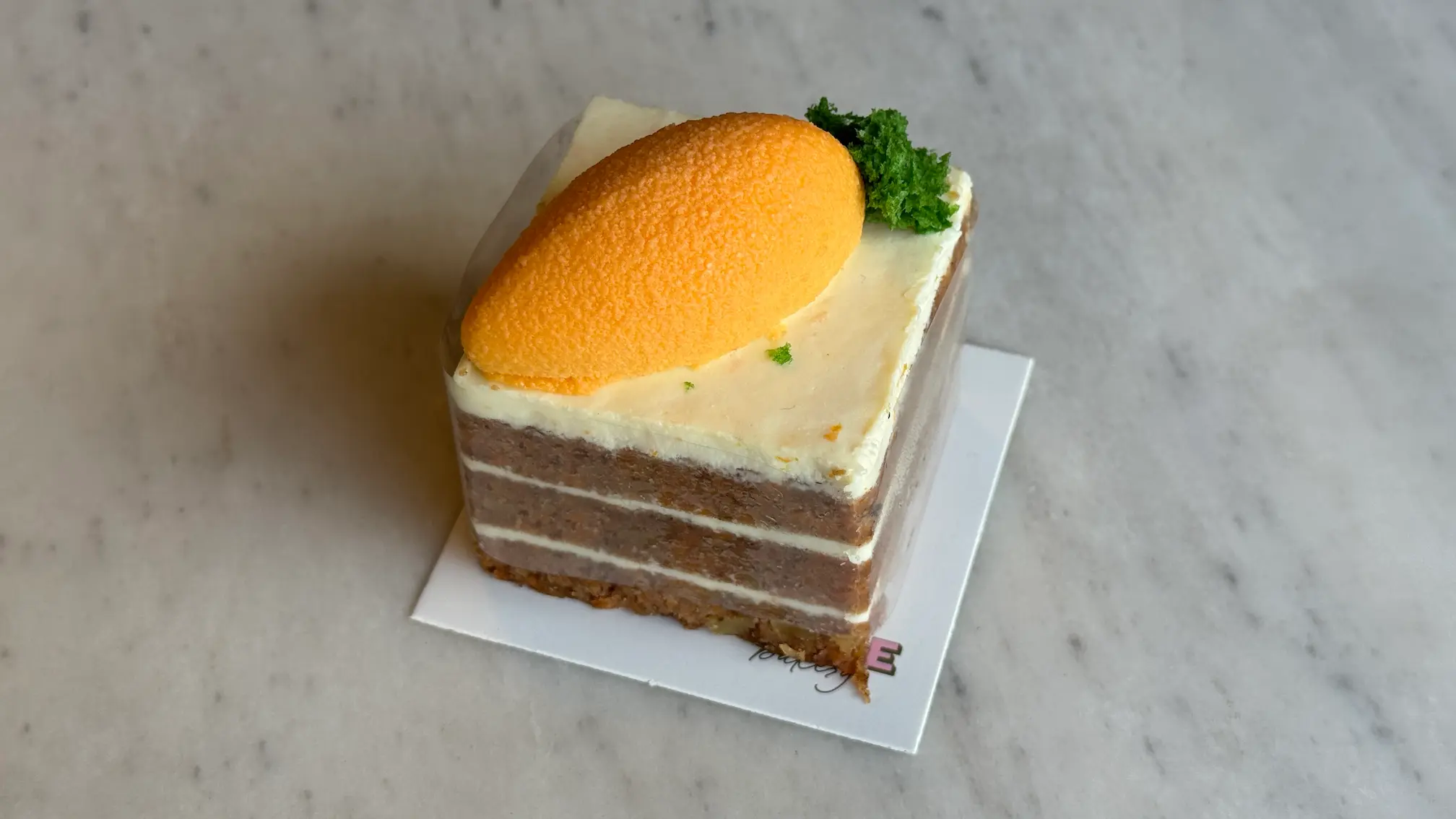 A person holds a cocktail glass containing a layered orange and yellow drink with a frothy top, set against a cozy bar background with warm lighting.