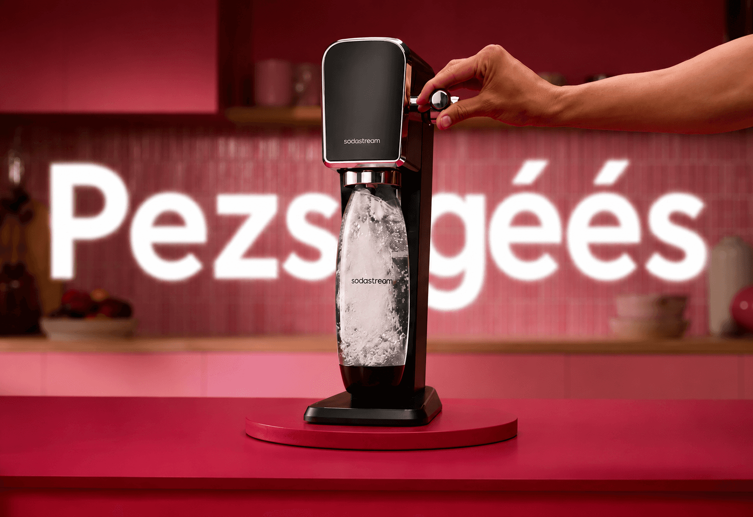 Szódagép használat közben, kéz működteti, háttérben felirat.