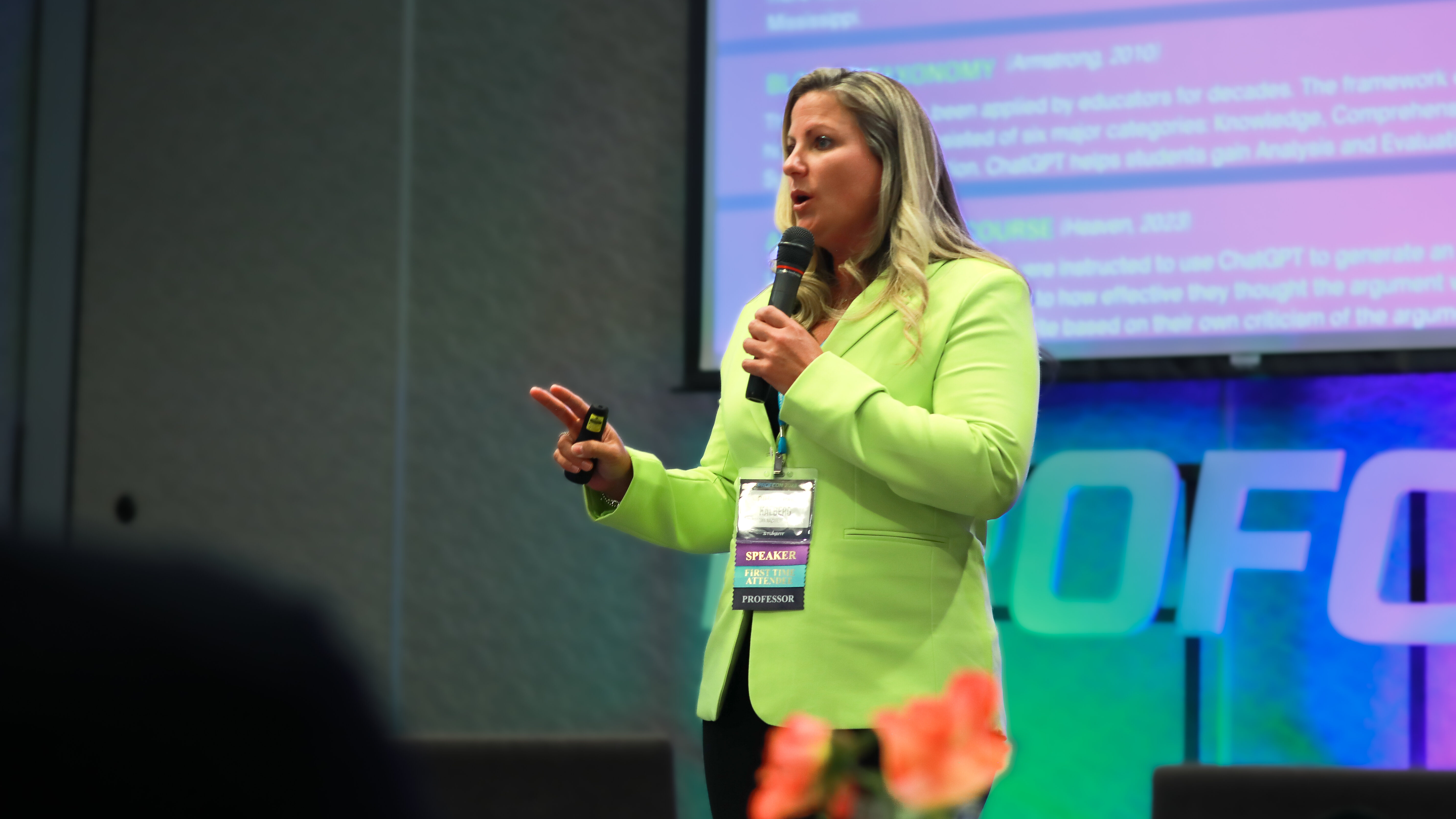 Female speaker in bright green blazer presents at PROFCON. She holds a microphone and clicker, gesturing with her hand. Her badge reads “SPEAKER – PROFESSOR.” A slide with text and citations is partially visible behind her.