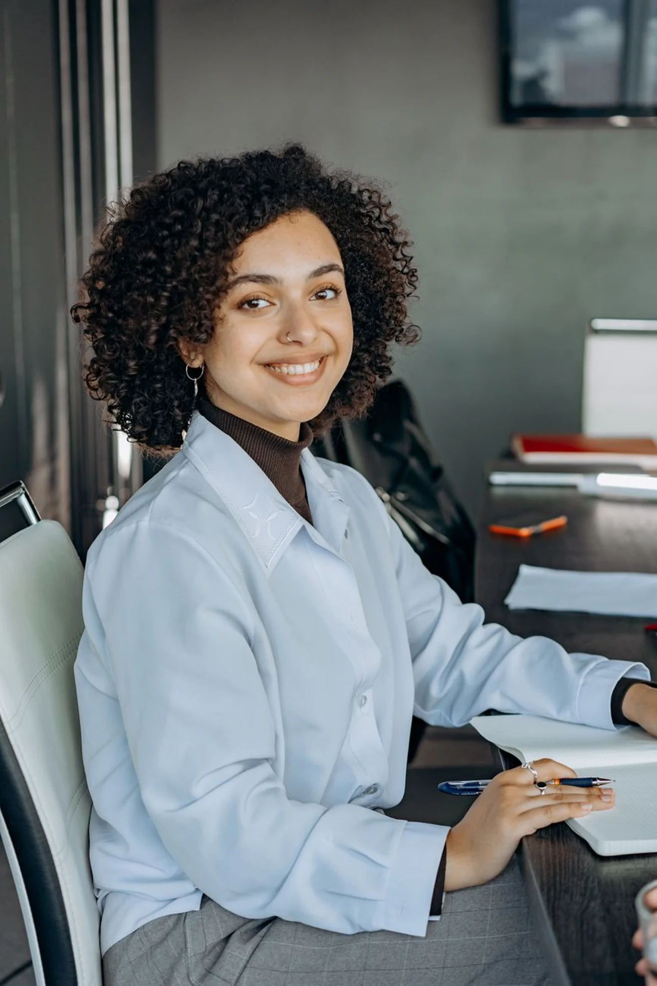 A portrait of a woman with short curly hair wearing a light blue button-down shirt, sitting at a conference table and smiling directly at the camera while holding a pen.